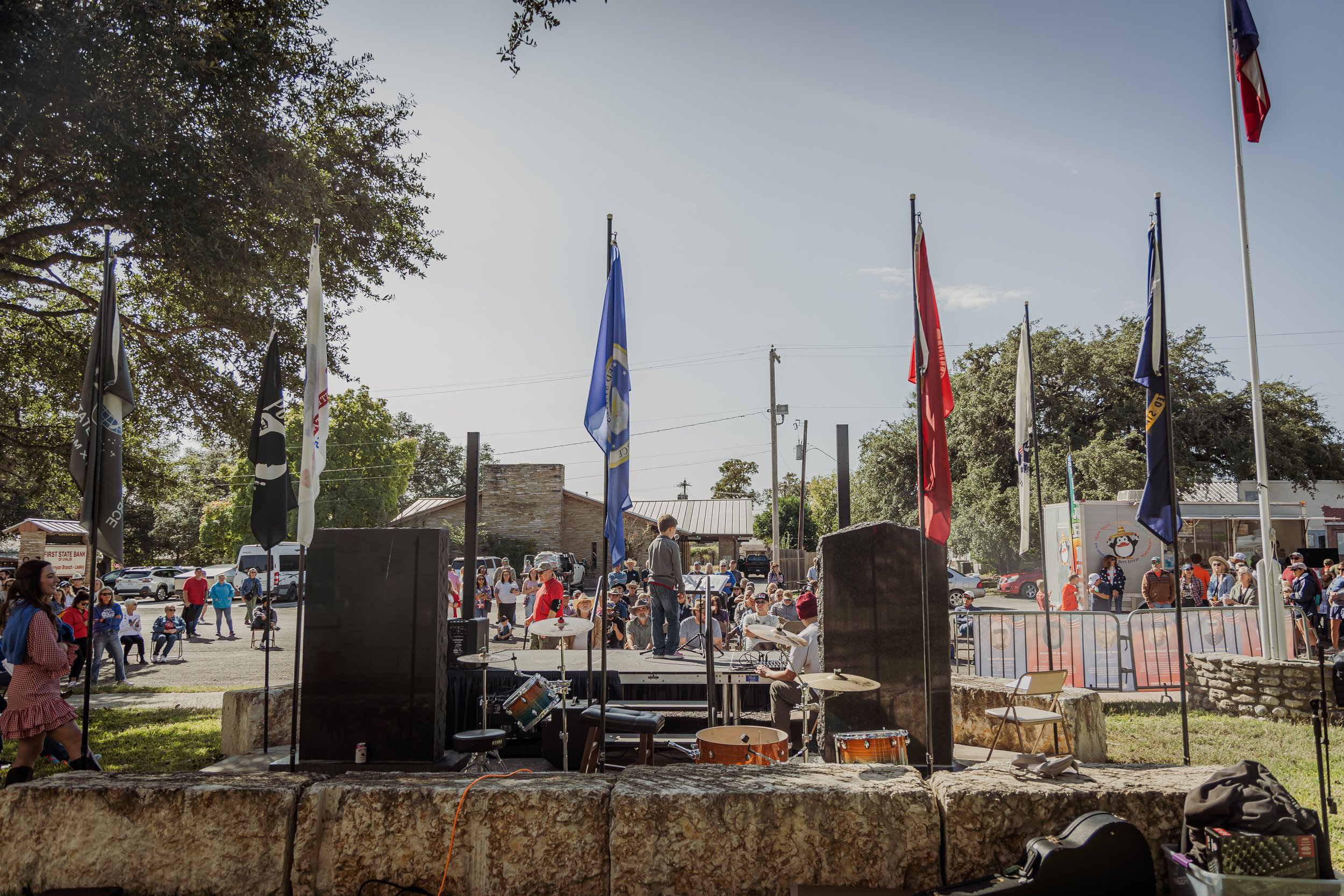 An outdoor event in a park with a stage set up in front of an audience, flags flying above, and a musician's drum set on stage. People are gathered around, some sitting and some walking, with trees and buildings in the background.