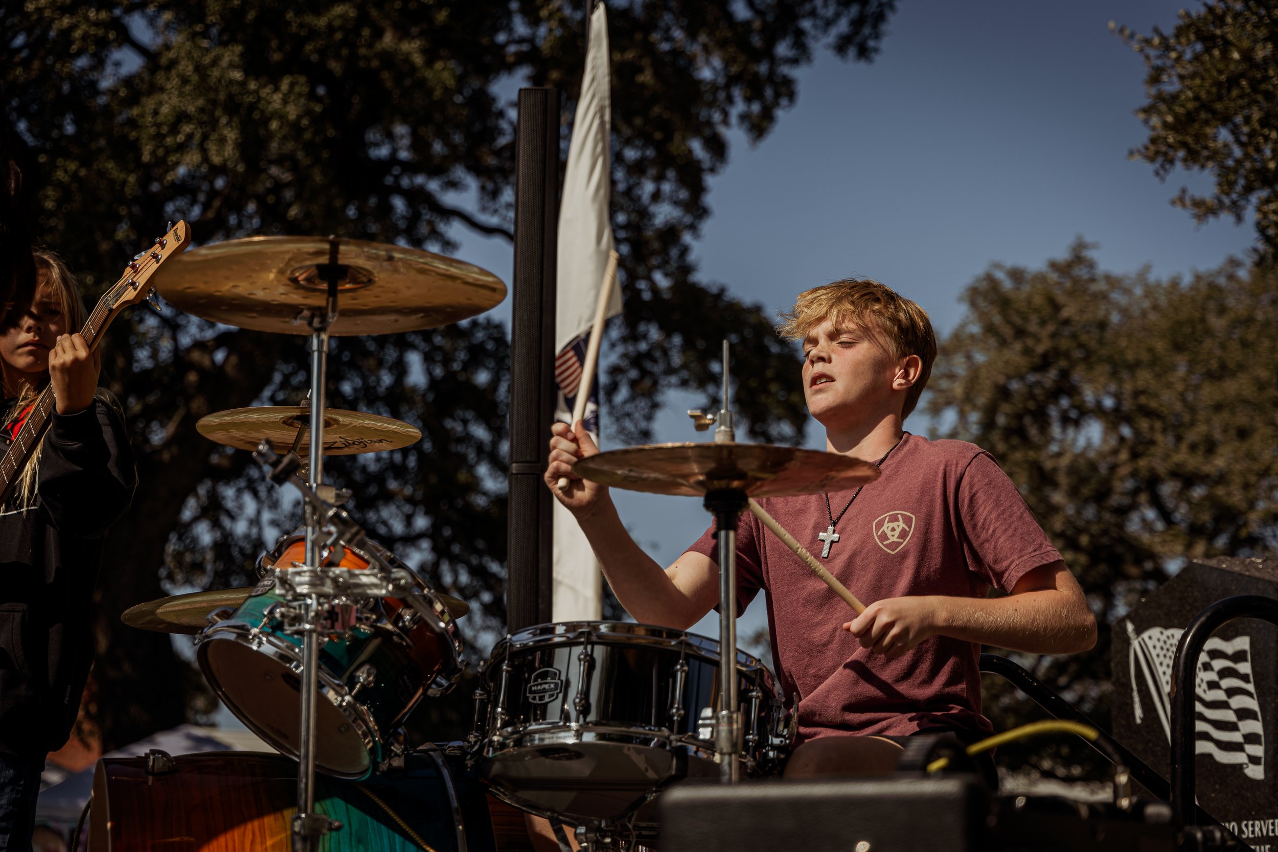 A young male drummer playing a drum set outdoors during daytime, with another person partially visible on the left holding a guitar. The drummer is wearing a maroon t-shirt and a cross necklace and appears focused. Large trees are in the background.