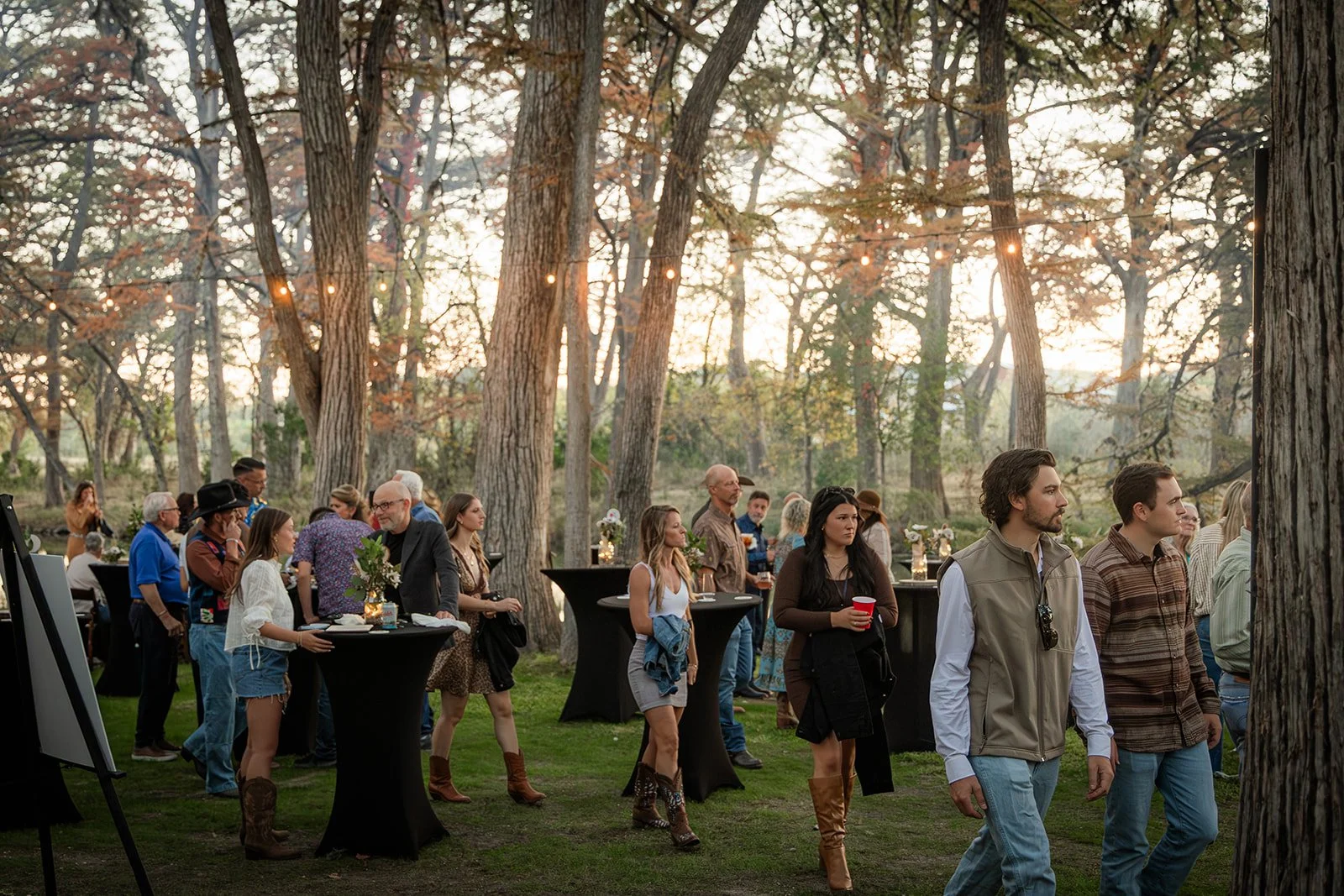 People gathered outdoors in a forest during sunset, standing around cocktail tables with floral centerpieces, dressed in casual and semi-formal attire, some holding drinks.