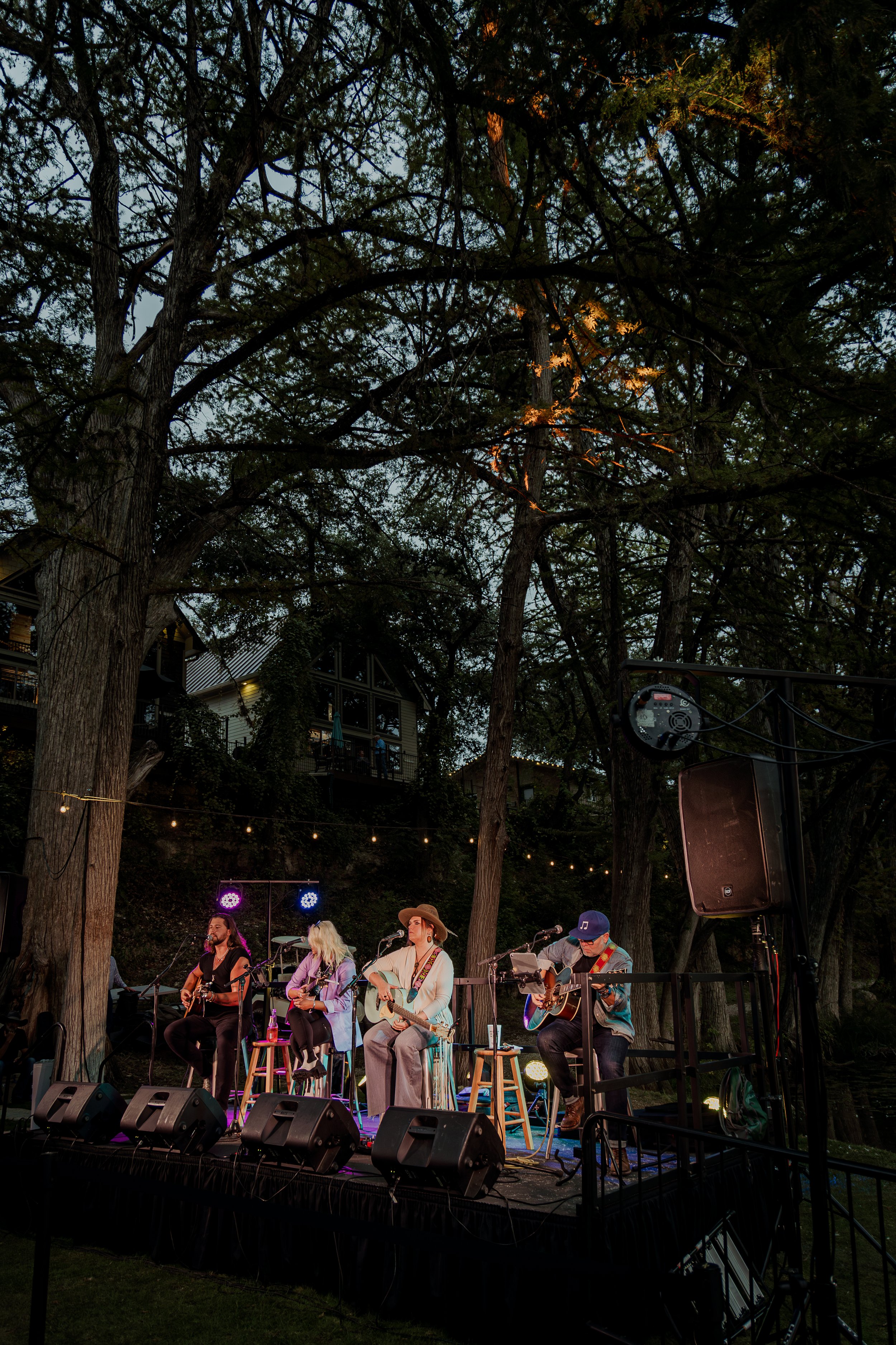 A live outdoor concert featuring four musicians performing on a stage under tall trees with houses in the background, stage lighting, and audio equipment.
