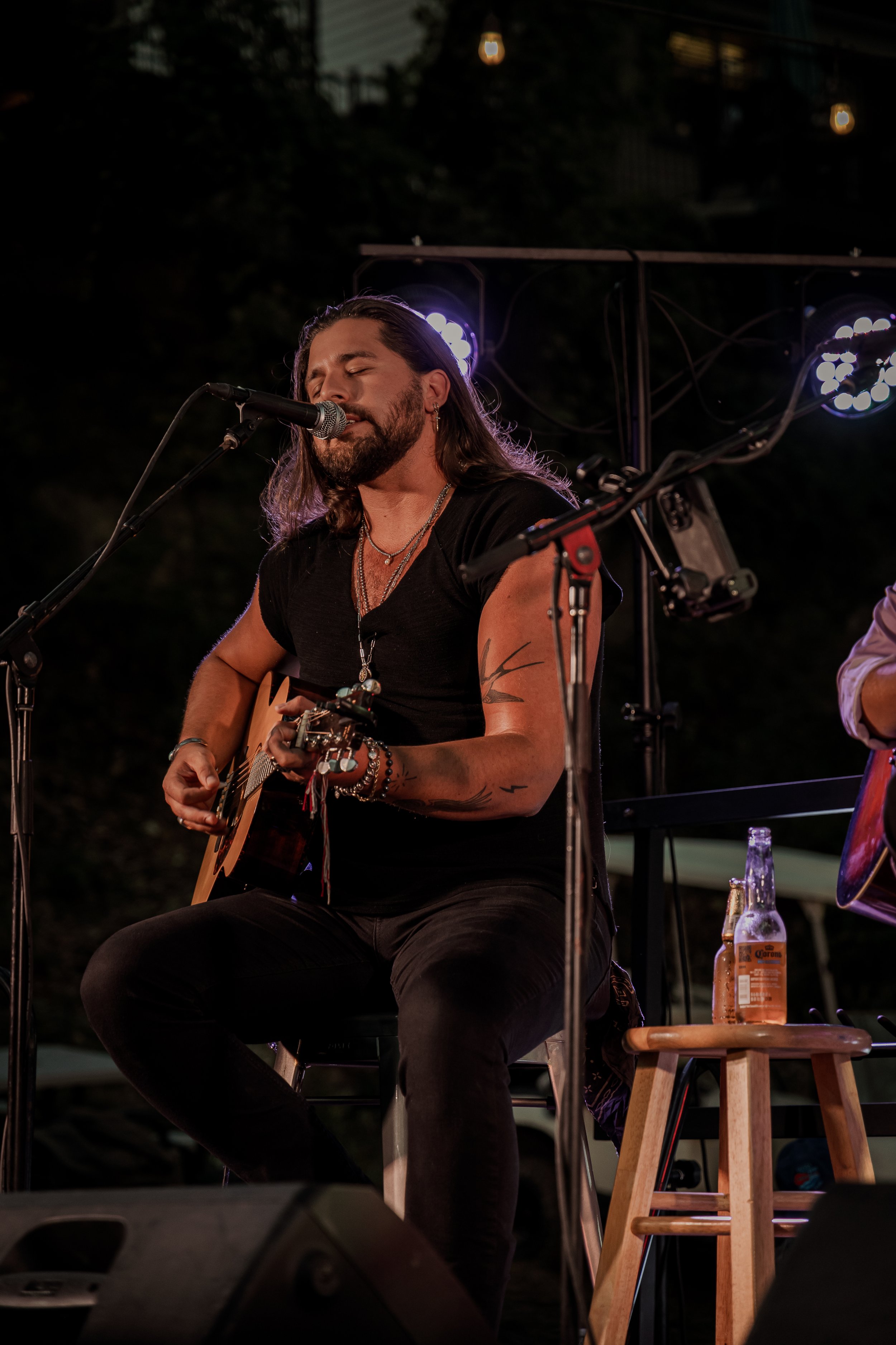 A man with long hair, beard, and tattoos, wearing a black V-neck T-shirt and jewelry, is performing on stage with a guitar, singing into a microphone at night. There are purple stage lights behind him and a Corona beer bottle on a small wooden stool