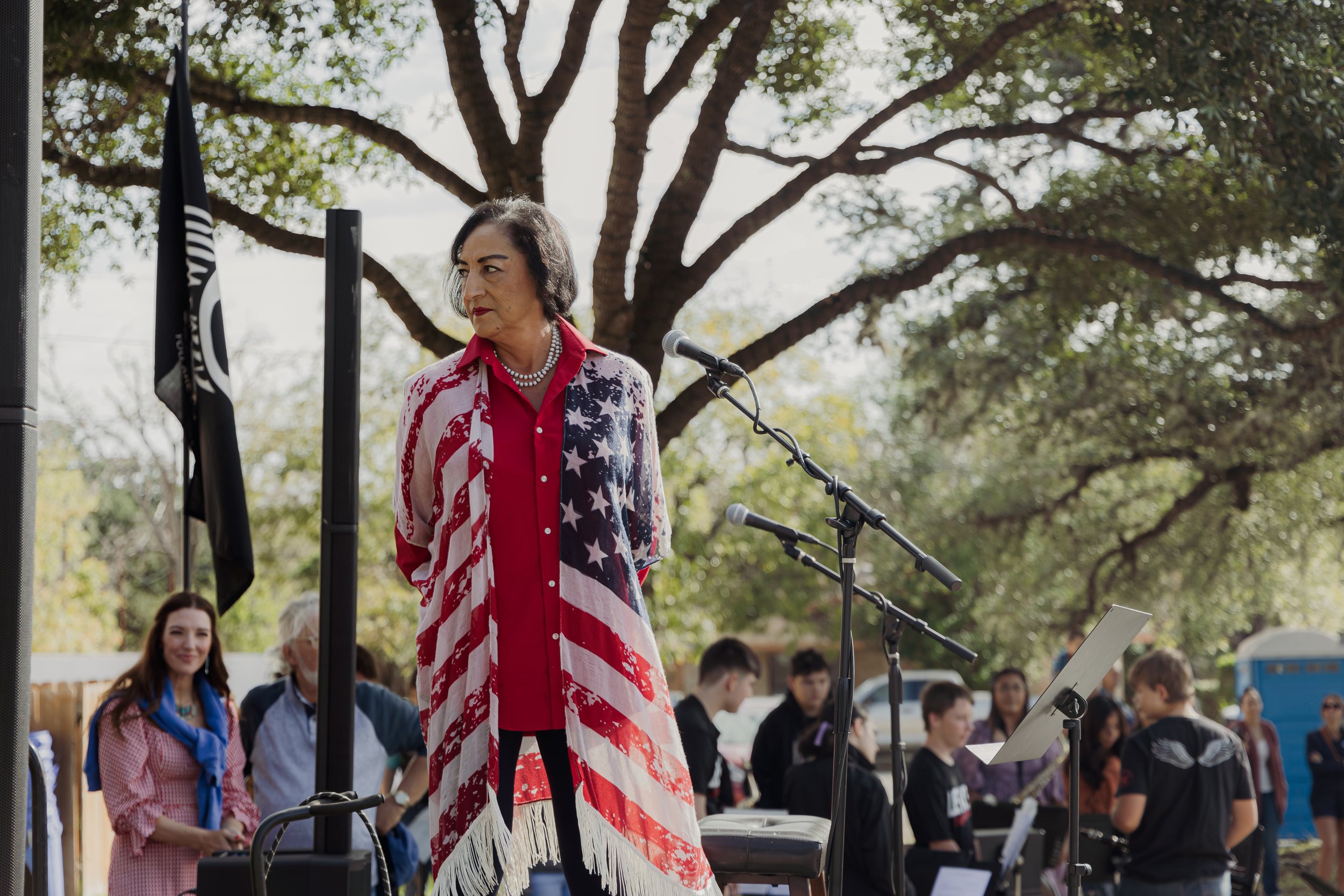 A woman standing on a stage or platform with a microphone, wearing a red shirt and a shawl with an American flag design. Behind her are musicians and people, with trees and a blue portable toilet in the background.