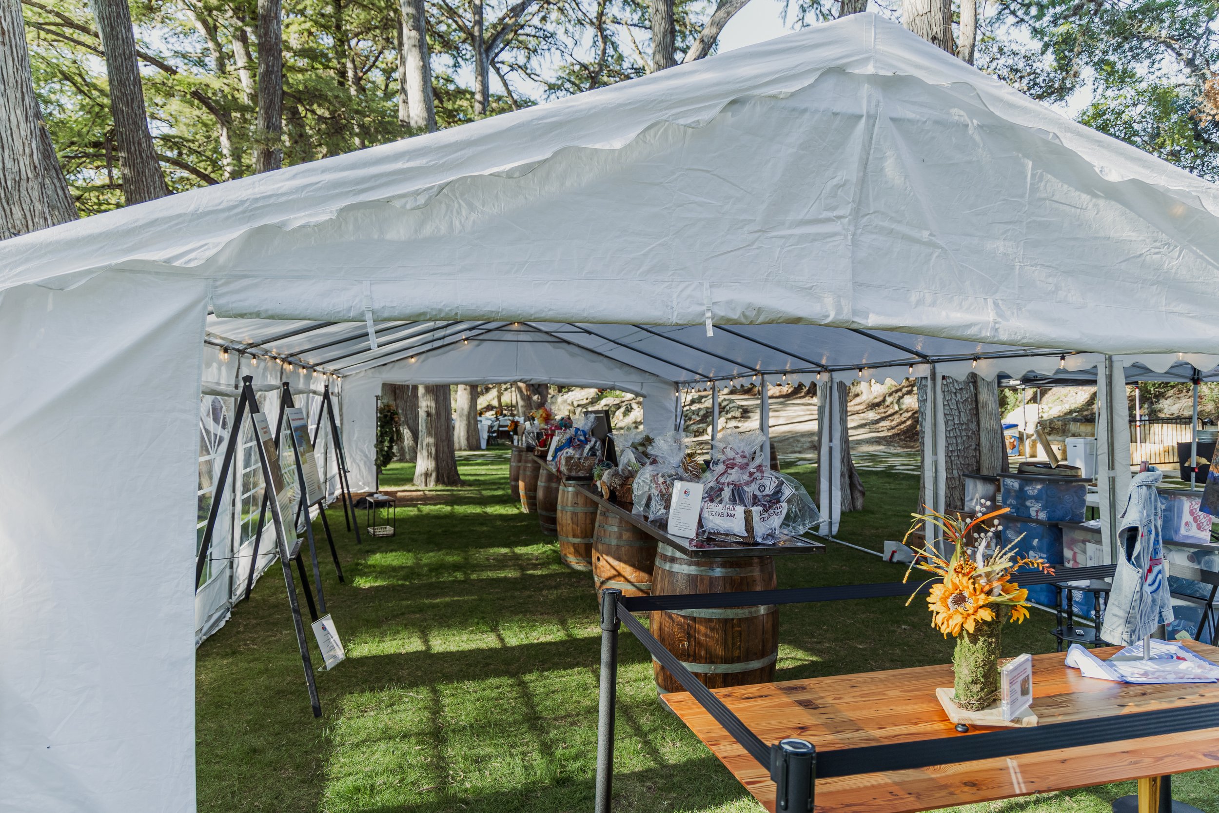 A white event tent set up outdoors in a grassy area, decorated with string lights and featuring wooden barrels with gift baskets, an array of framed photos on easels, and a wooden table with a flower arrangement.