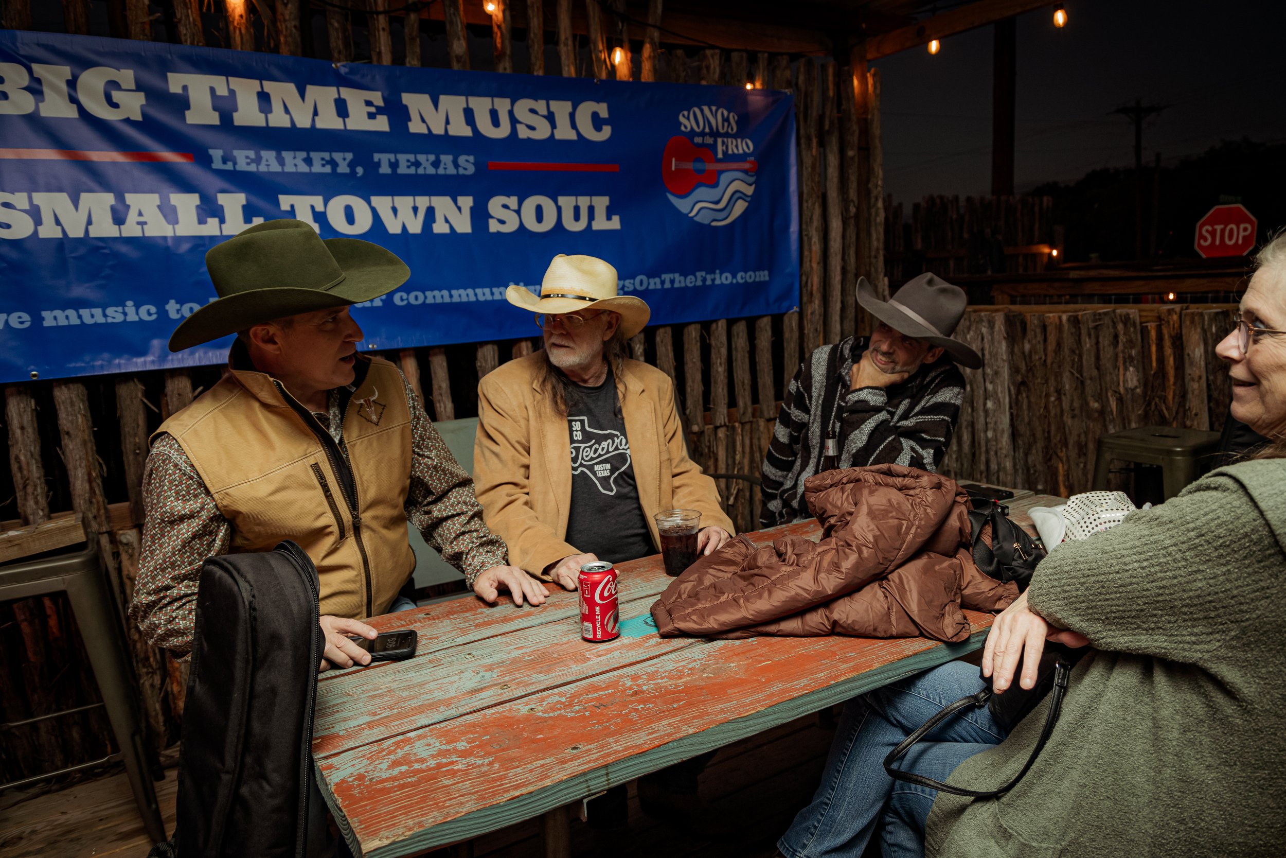 Four people sitting at a rustic outdoor table, engaged in conversation. The background features a blue banner with white text that reads 'Big Time Music, Small Town Soul,' and a logo for 'Songs of the Frio.' One person wears a cowboy hat and vest, an