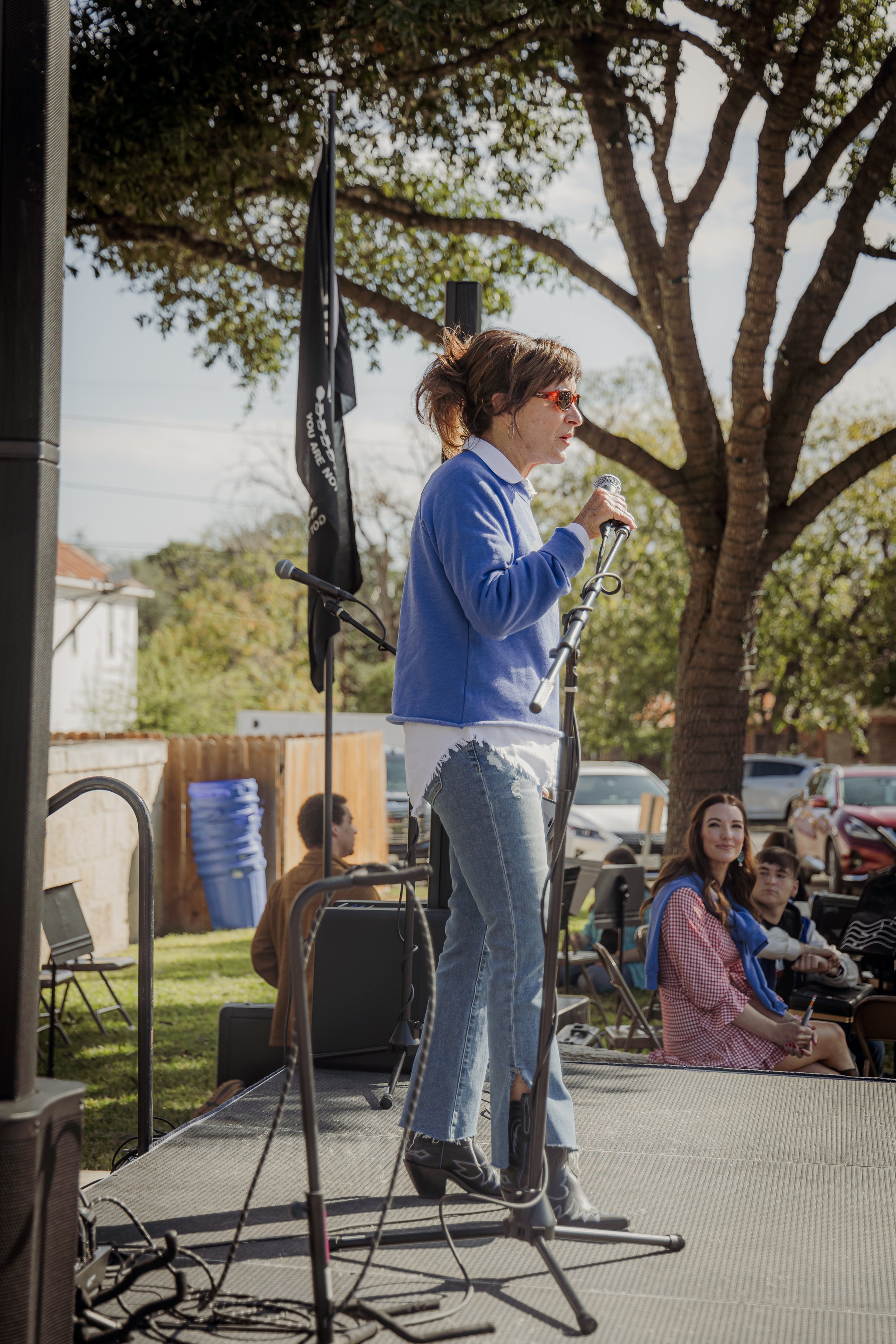 A woman wearing sunglasses and a blue jacket speaks into a microphone on an outdoor stage, with a group of people seated and standing behind her under a large tree.