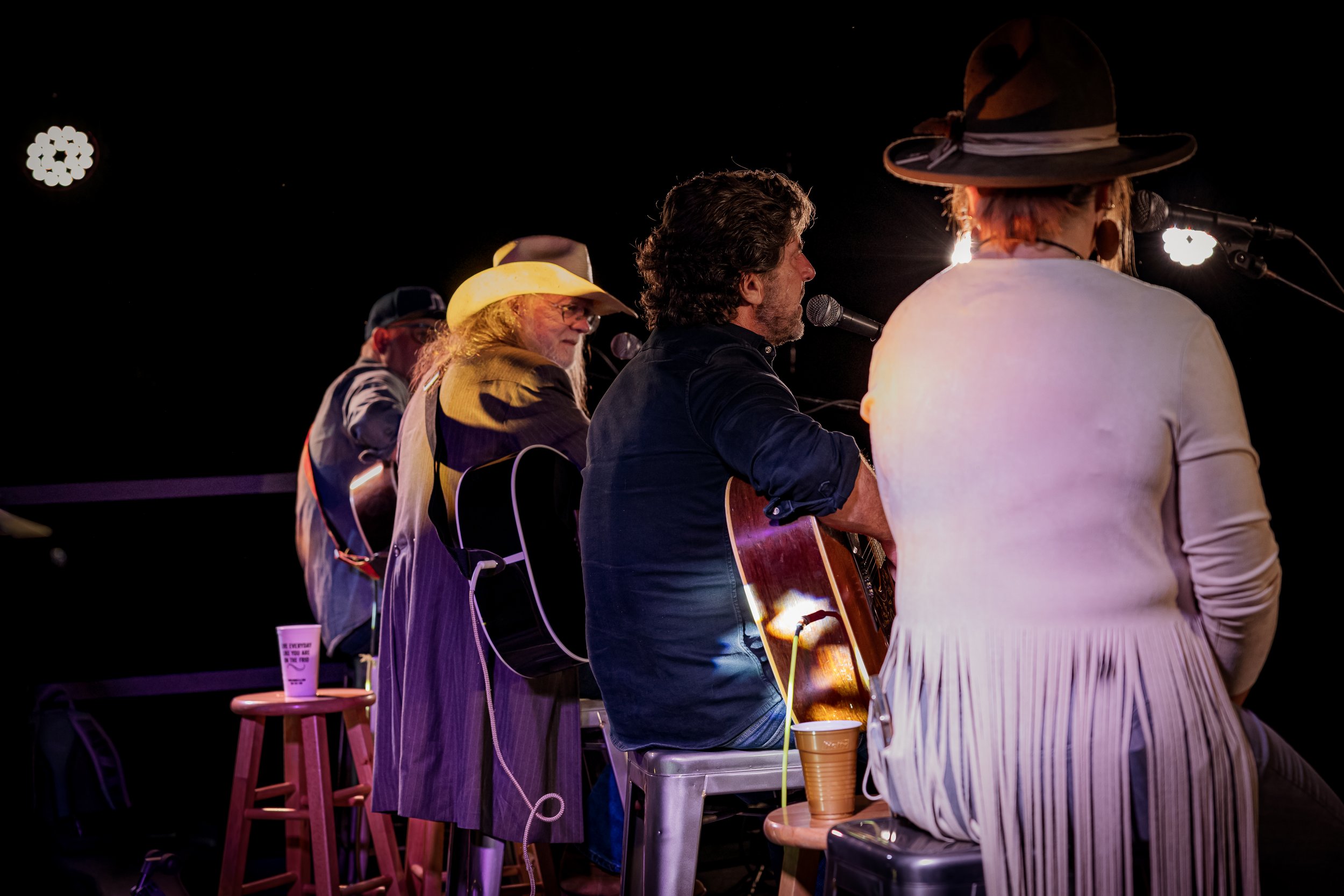 Four musicians playing guitars and singing on stage under stage lights, with one person in foreground wearing a large hat and another with a cowboy hat, seated on stools and a bench.