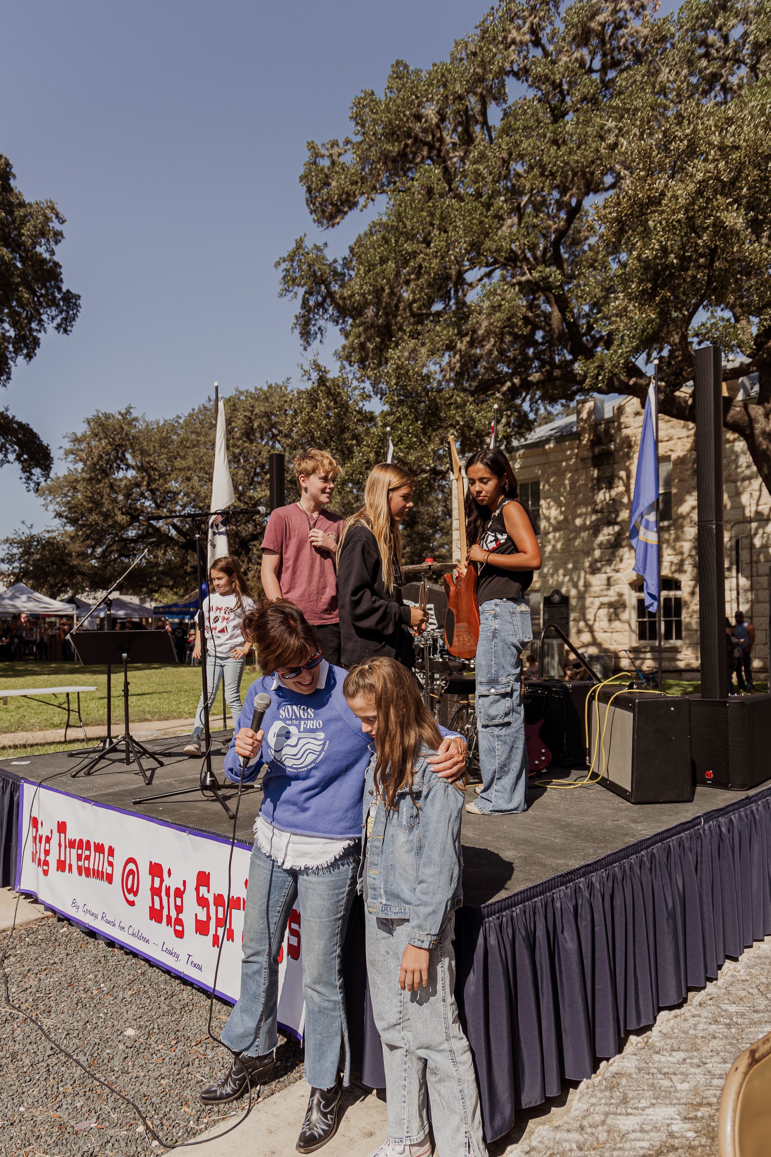 A group of children and a woman with a microphone, on an outdoor stage, engaging in a musical performance or activity at an event, with tents and trees in the background.
