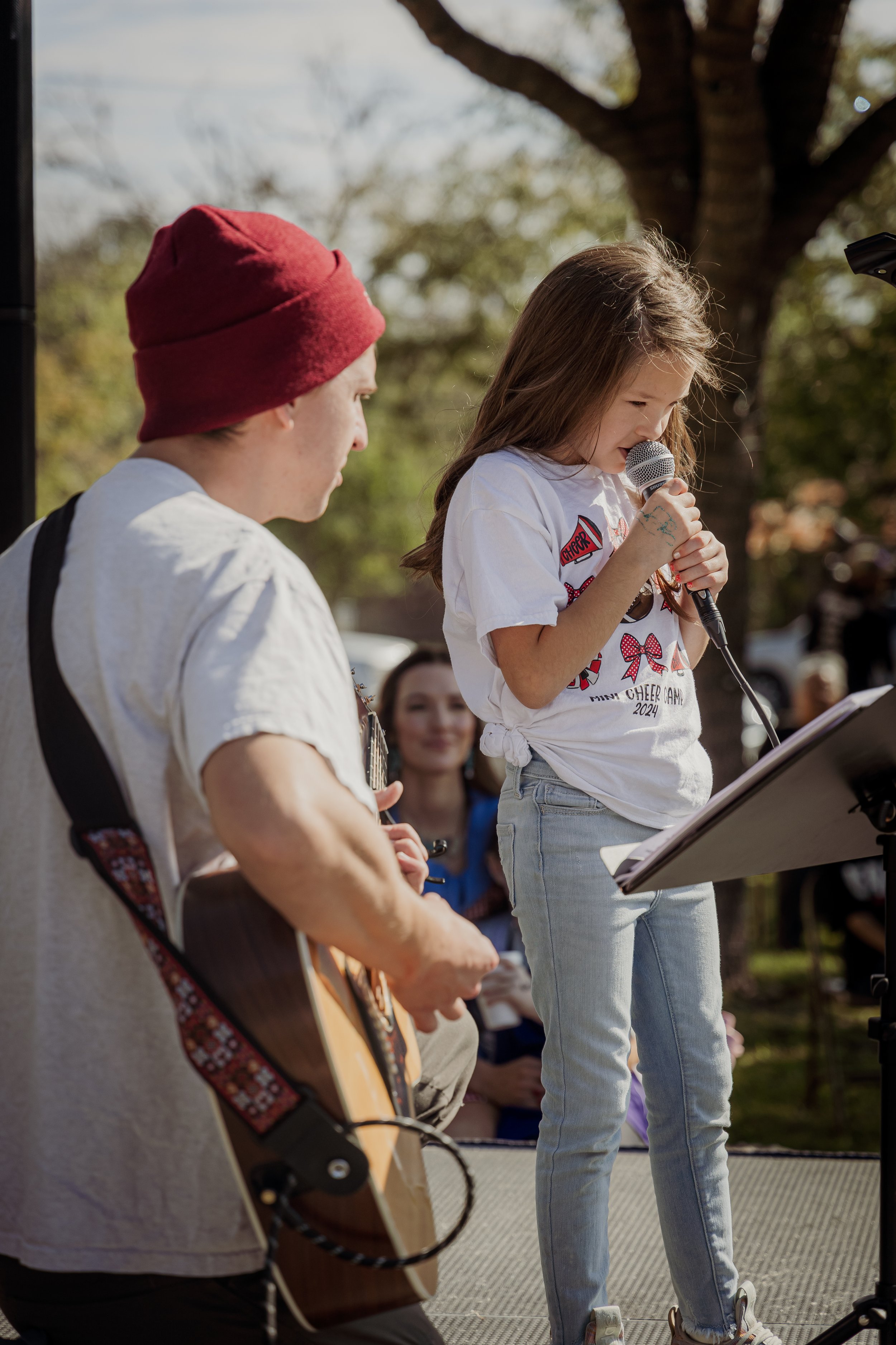 A young girl with long hair holds a microphone and sings while standing on a stage outdoors, accompanied by a guy playing an acoustic guitar. An audience is visible in the background under a tree on a sunny day.