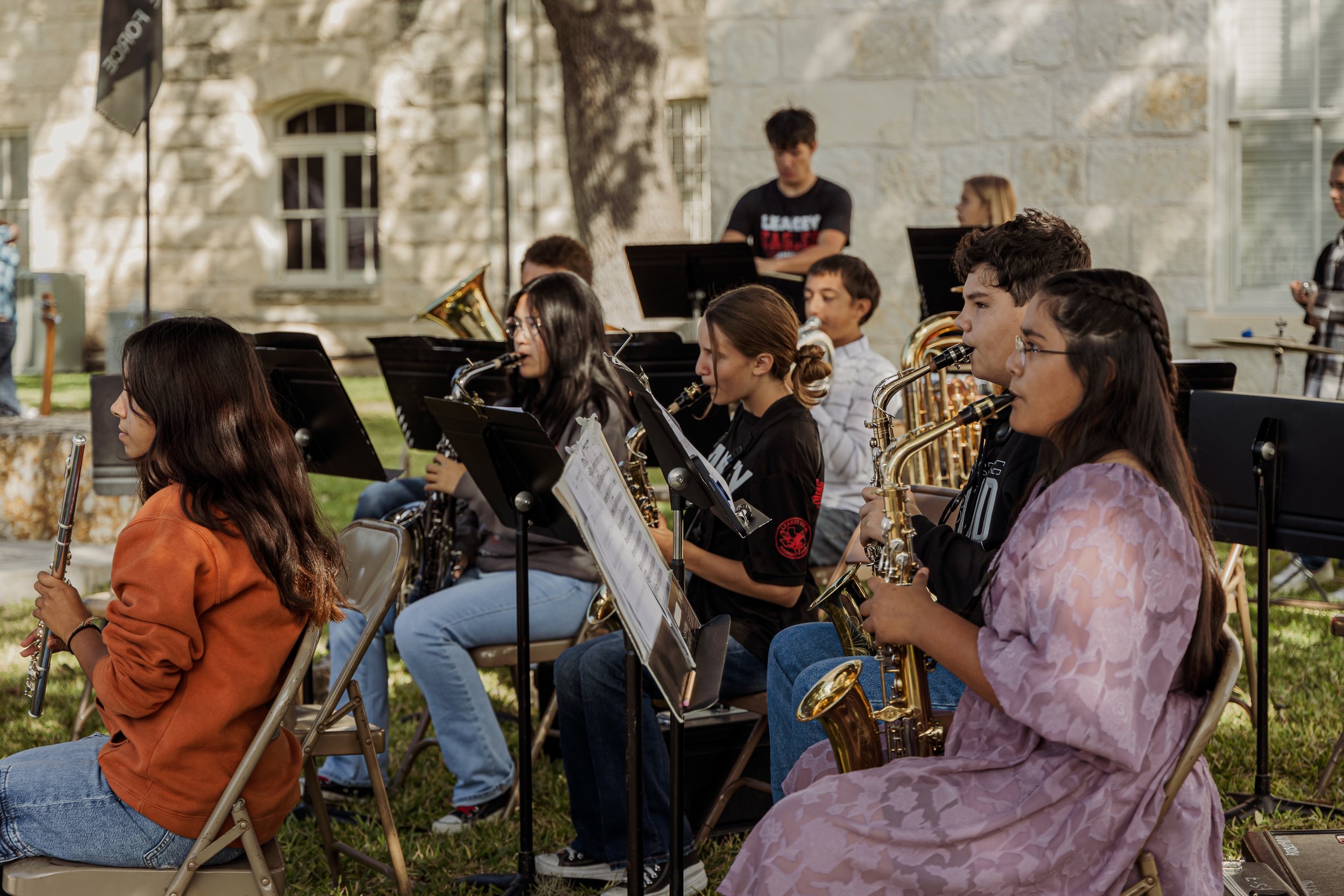 Group of young musicians playing wind instruments outdoors, sitting on chairs with music stands, in front of a stone building with windows and trees.