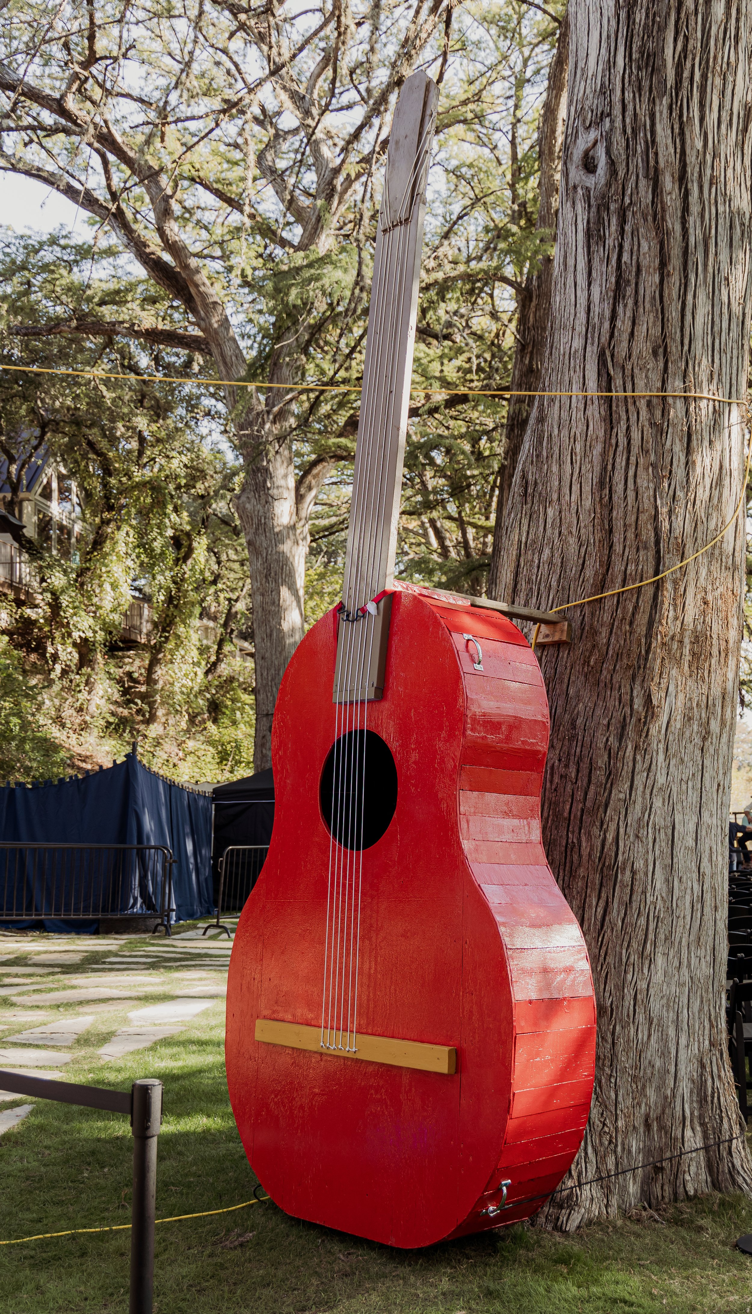 A guitar-shaped house with a red body and a neck leaning against a large tree outdoors.