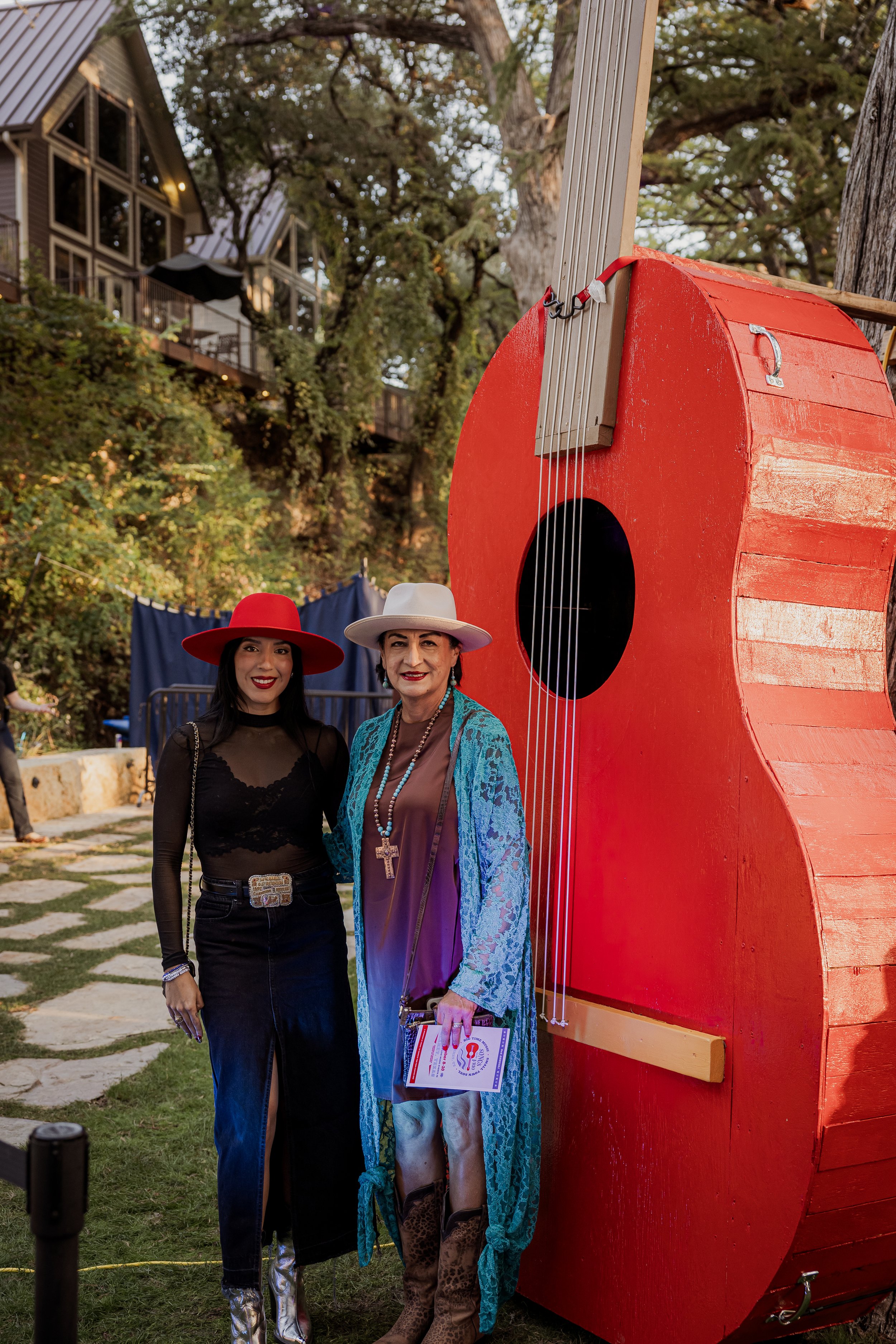 Two women wearing colorful hats and stylish outfits, standing next to a large red guitar-shaped sculpture outdoors with trees and houses in the background.