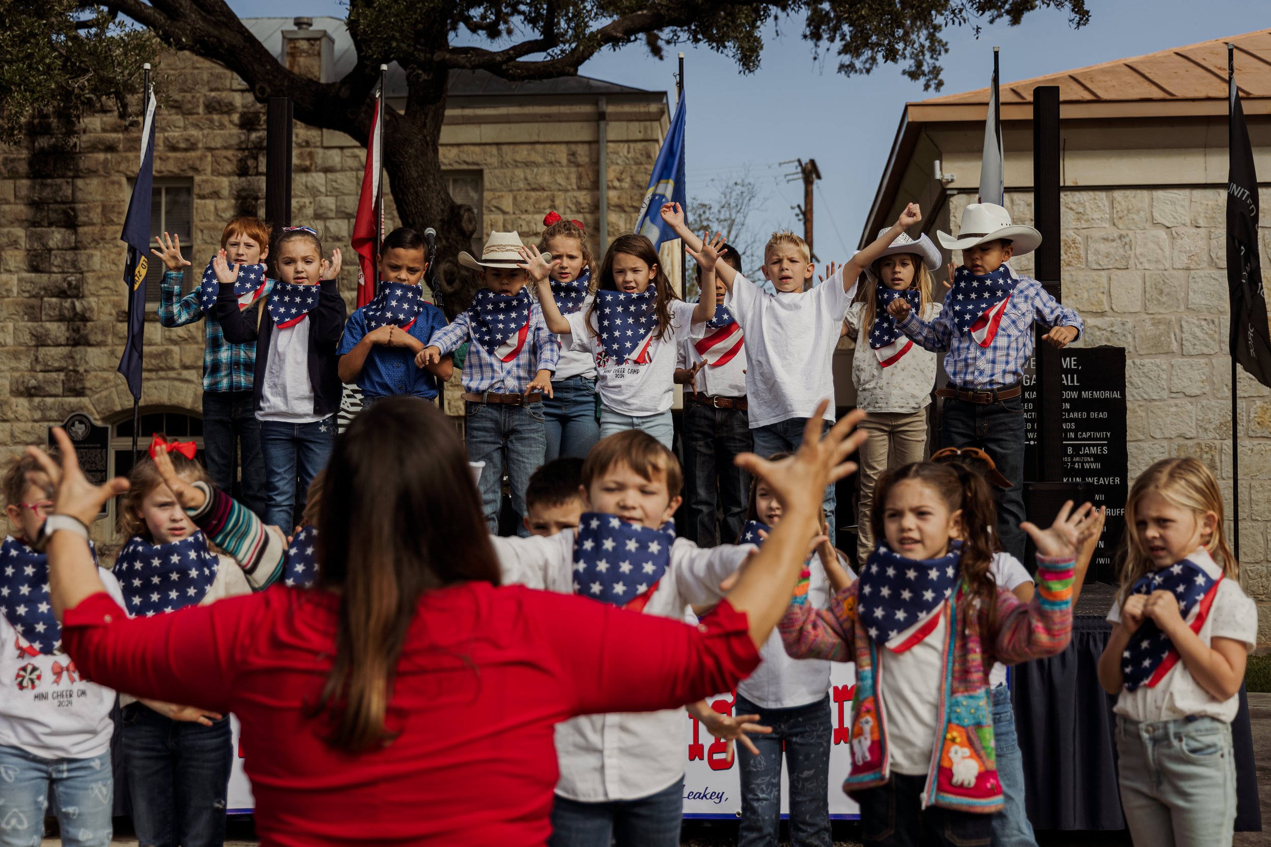 Children performing on an outdoor stage with patriotic themed bandanas and attire, directed by an adult woman in a red shirt, during a daytime celebration.
