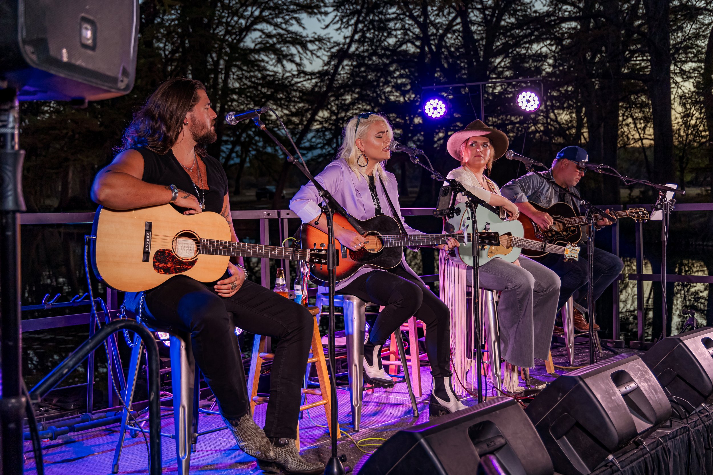 Four musicians performing on an outdoor stage during dusk, each sitting on stools, playing acoustic guitars. They are under purple stage lights with trees in the background.