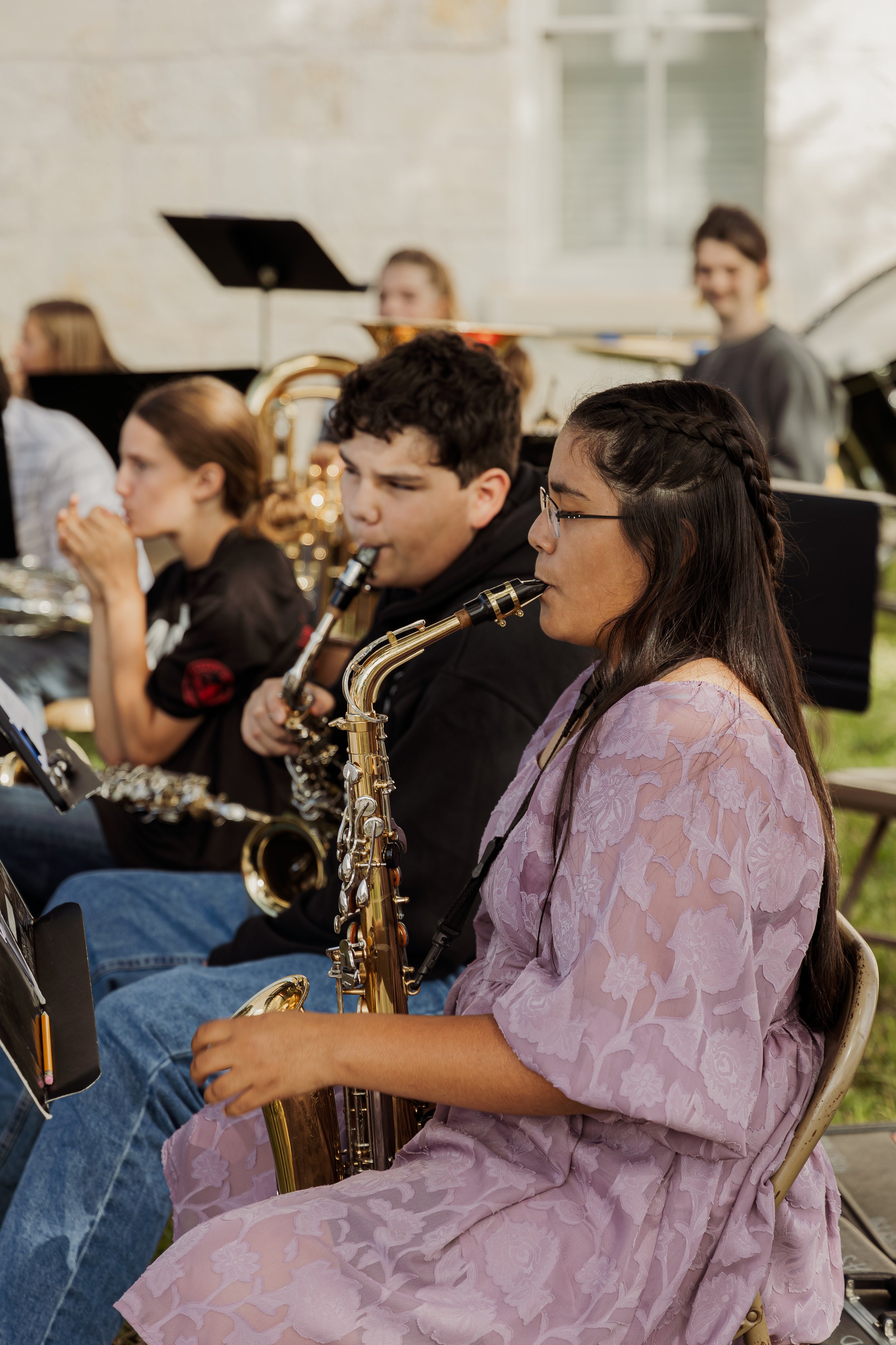 Young woman sitting and playing the saxophone during a band rehearsal outdoors with other musicians in the background.
