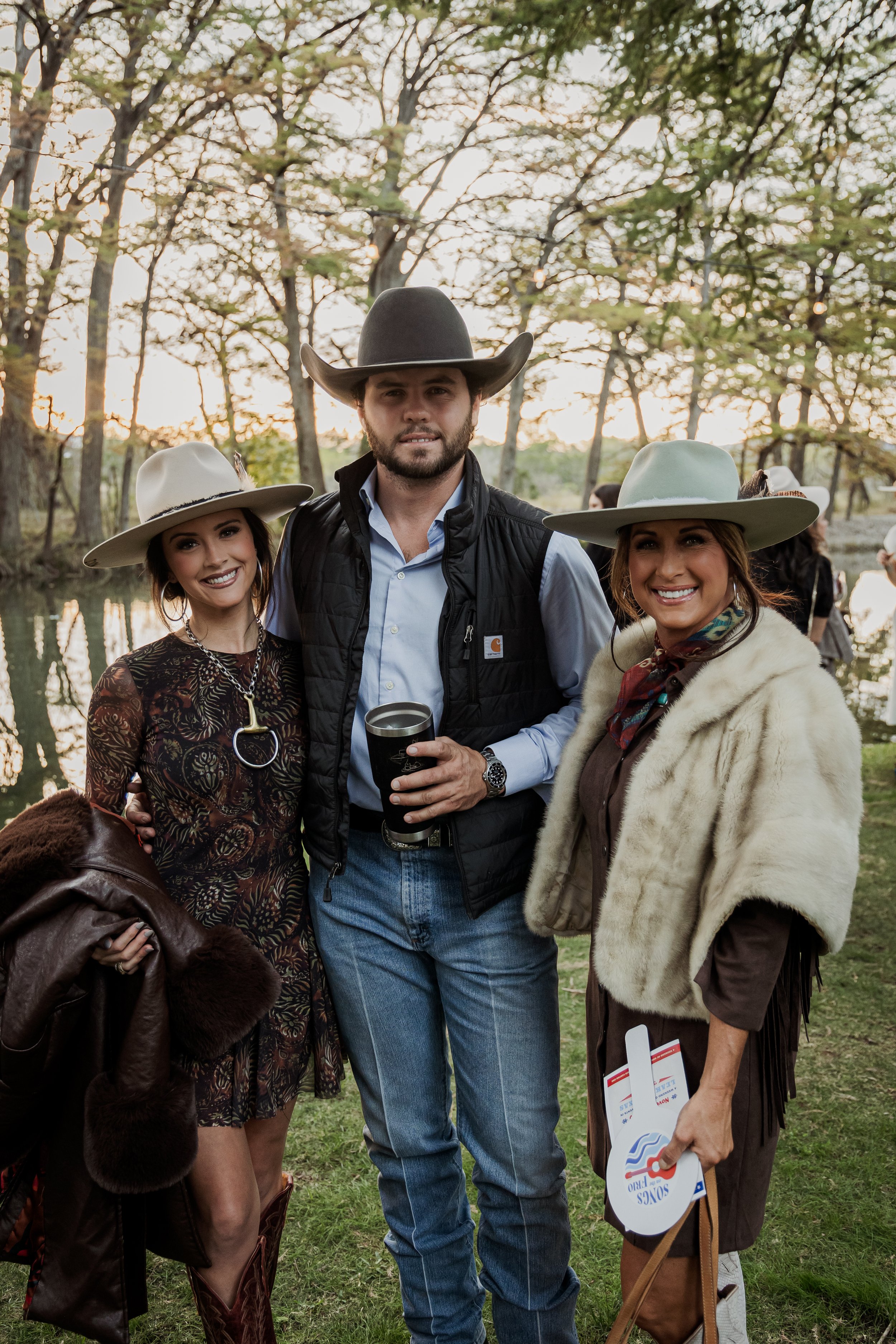 Three people dressed in western attire standing outdoors near a body of water with trees in the background during sunset