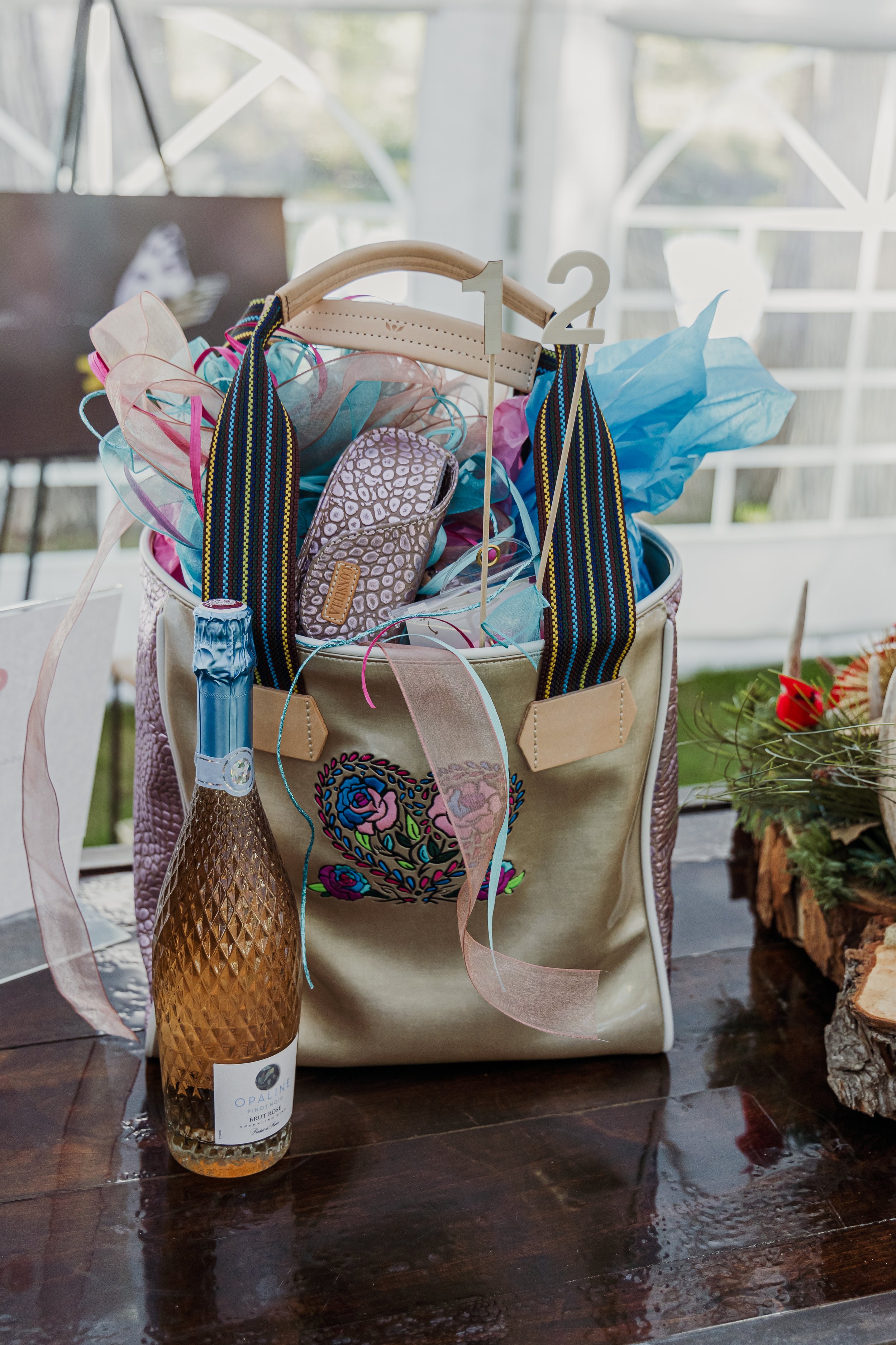 A gift bag with a floral embroidery design contains colorful tissue paper, a patterned eyeglass case, and a striped tote handle. There is a bottle of sparkling wine labeled 'Opaline' next to the bag, on a dark wooden table.
