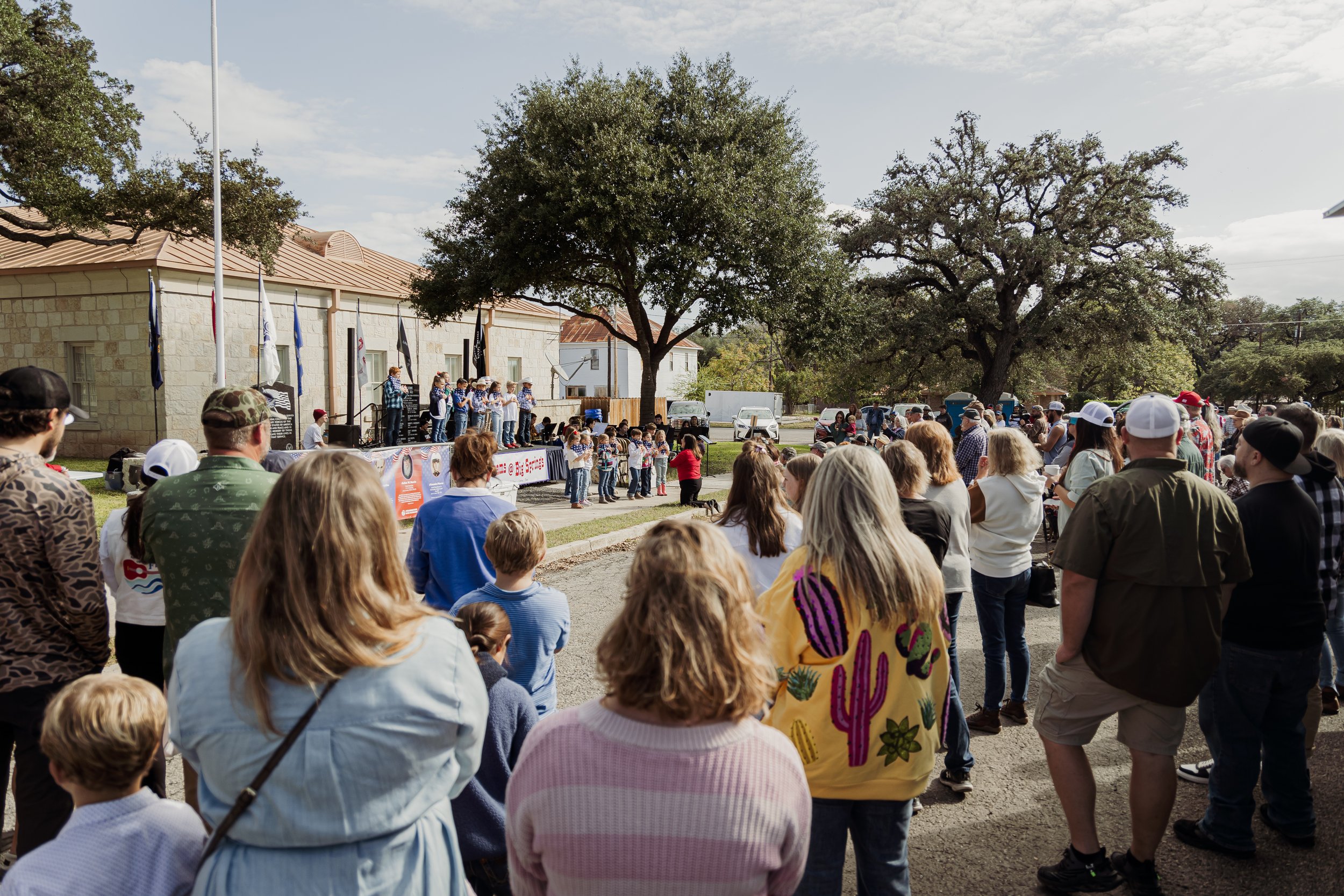 A crowd of people gathered outdoors watching children perform on a stage in front of a historic building, with large trees and a partly cloudy sky in the background.