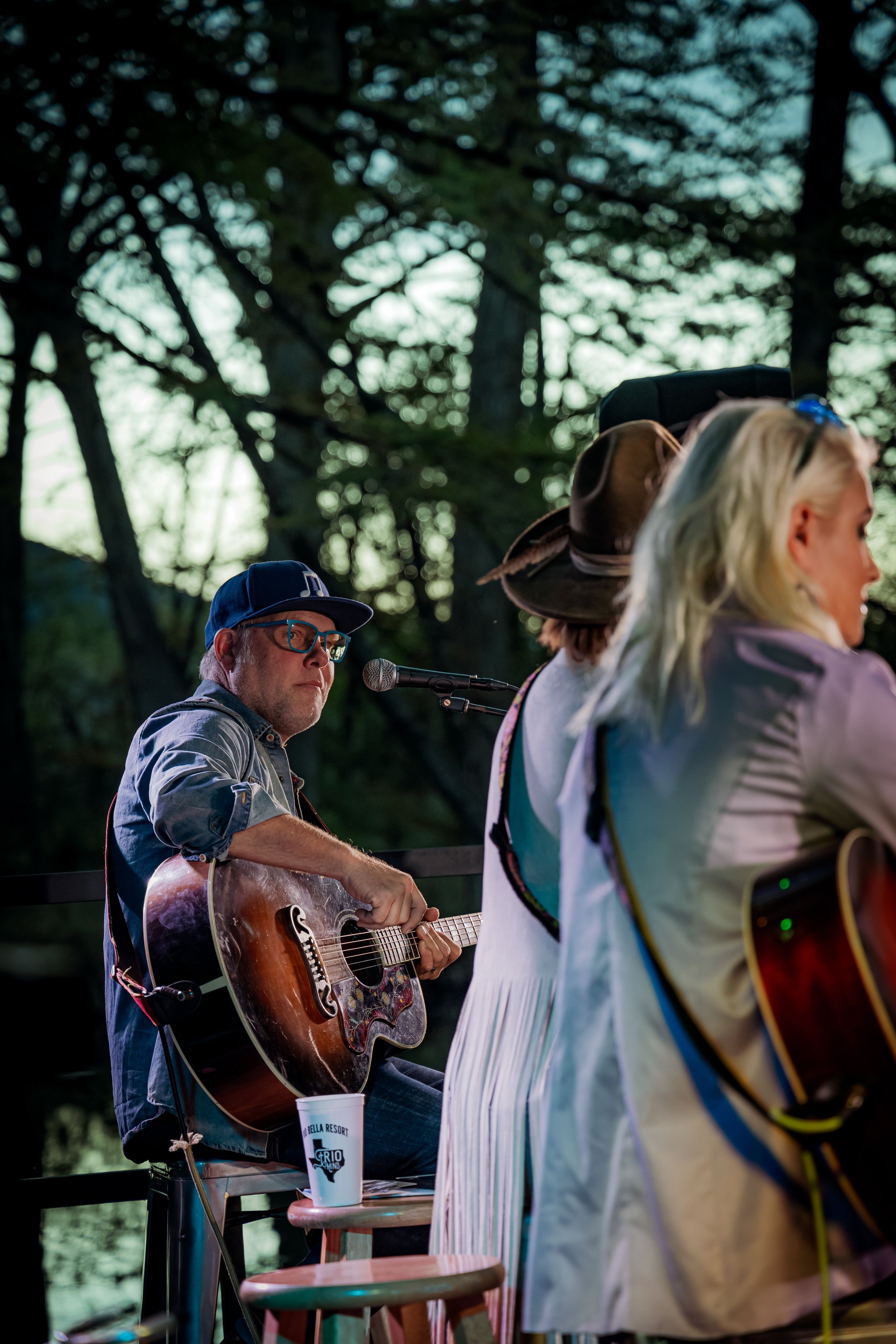 A man wearing glasses and a blue cap playing an acoustic guitar during an outdoor performance, with a microphone nearby and trees in the background.