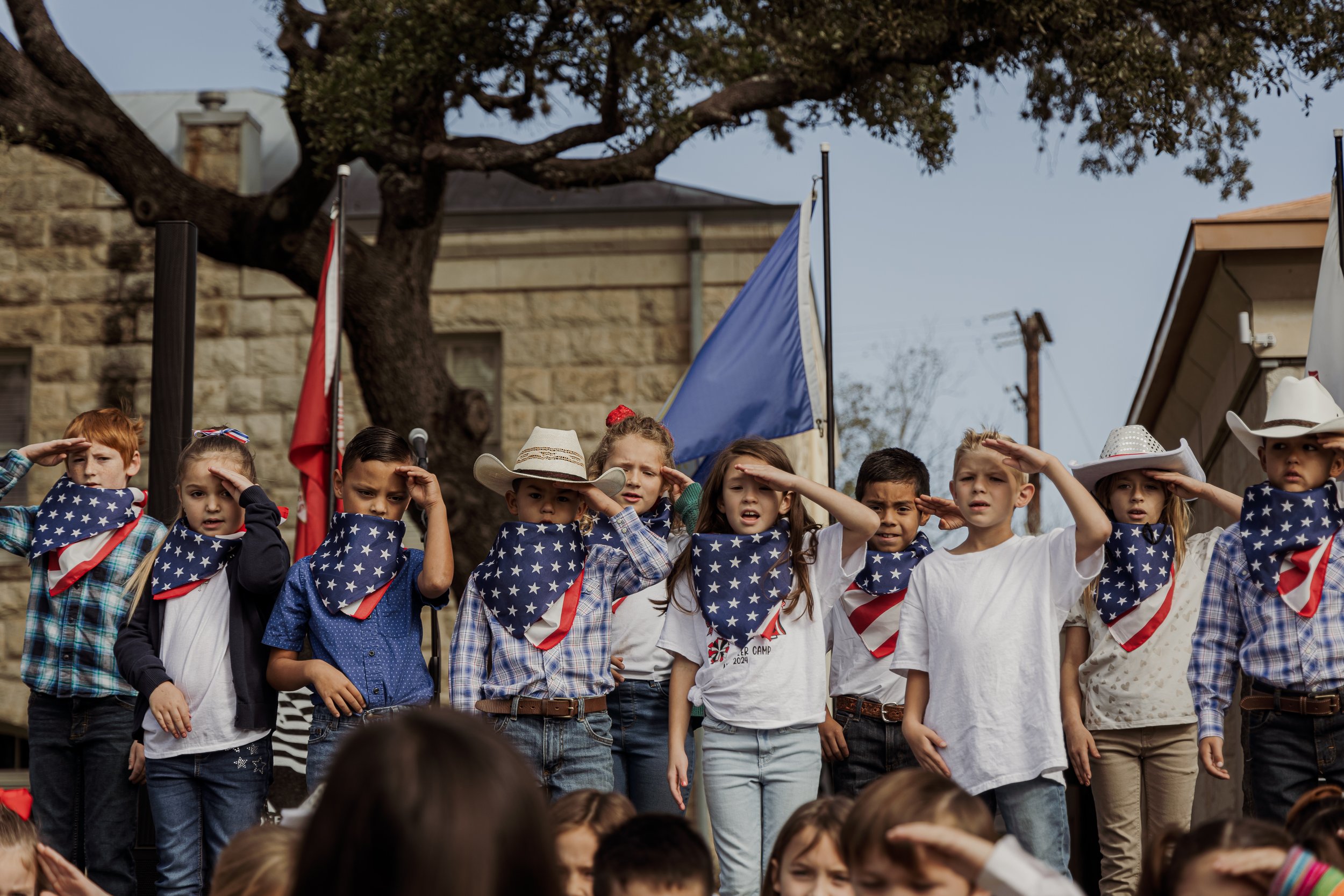 Children dressed in patriotic outfits, saluting during a parade or ceremony, with American flags and banners in the background.