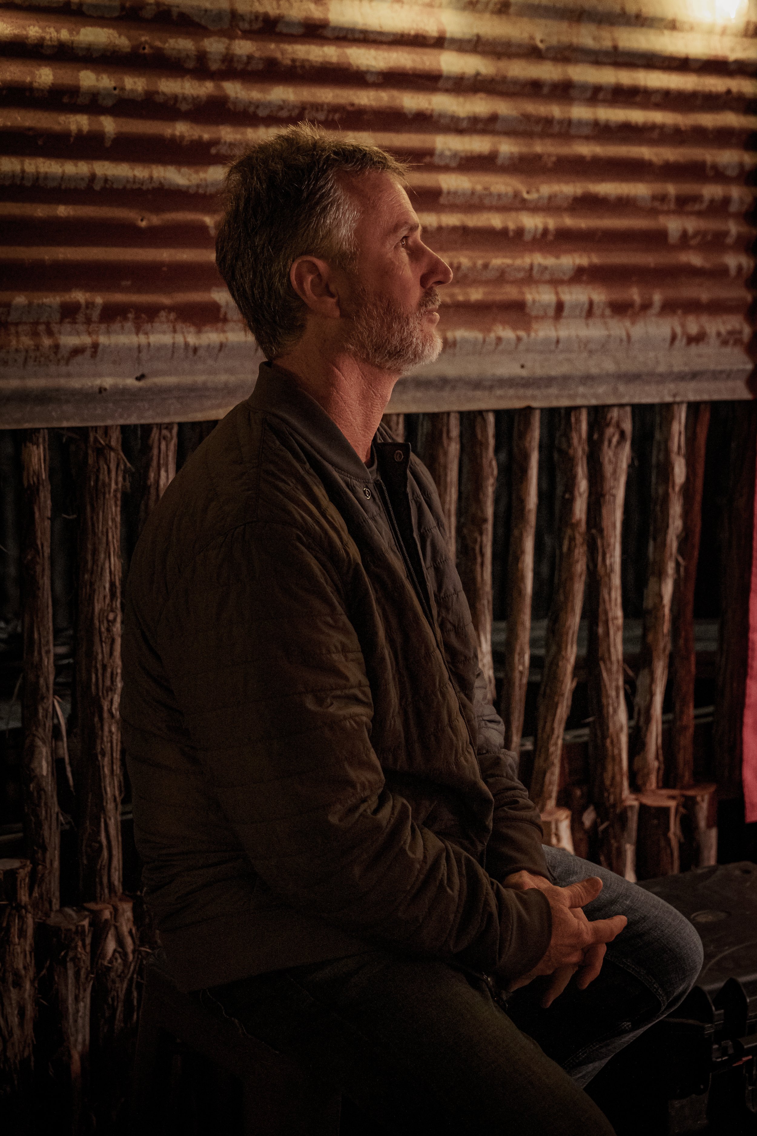 Side profile of a middle-aged man with gray hair and beard sitting indoors, wearing a brown jacket, in front of a rustic wooden wall.