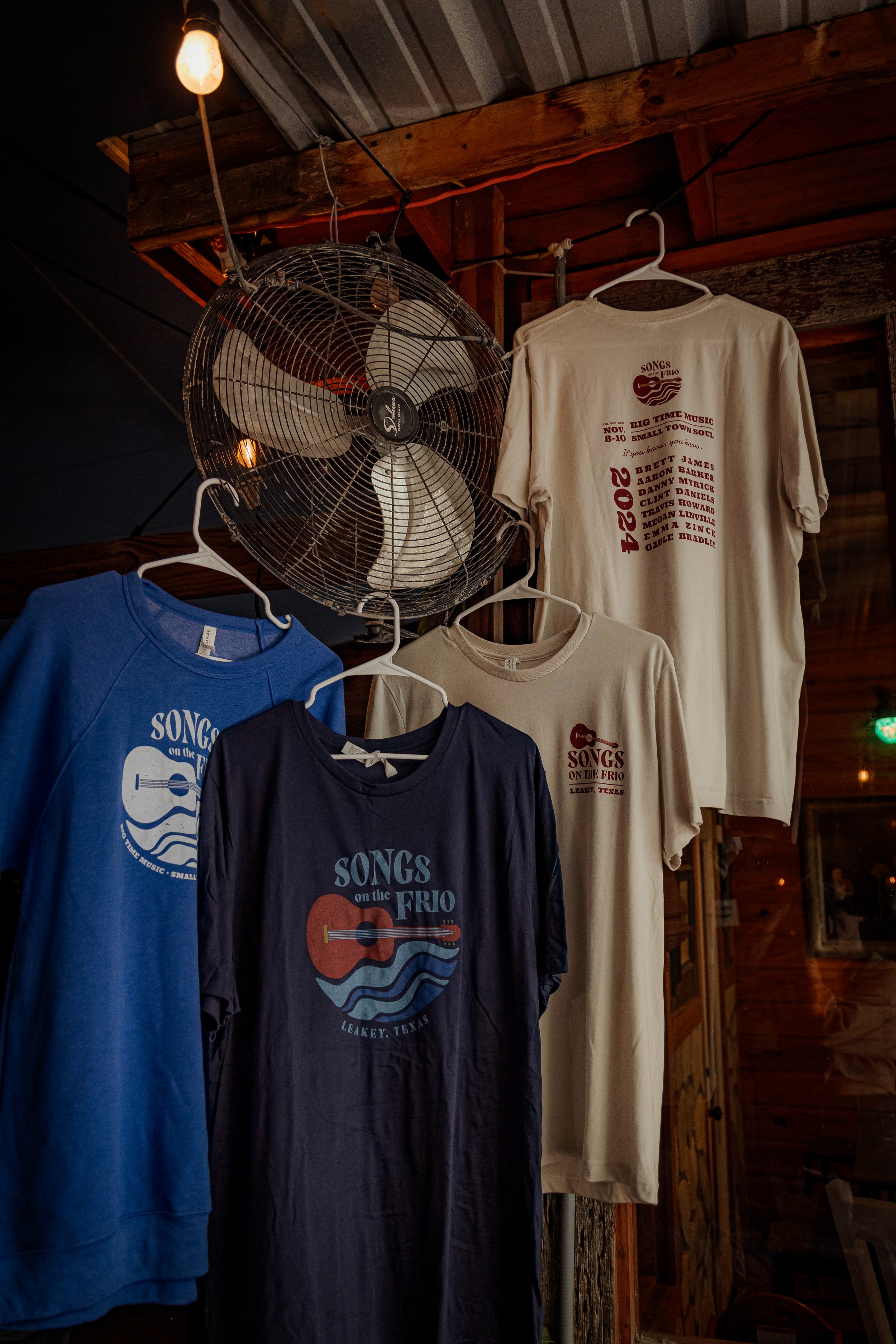 T-shirts hanging on a wooden wall with a vintage-style ceiling fan above, advertising a 2024 music event called Songs on the Frio in Leakey, Texas.