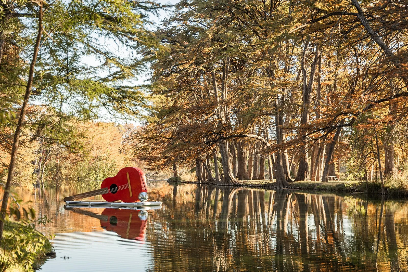 A red guitar figurine floats on a small raft on a calm river surrounded by trees with autumn foliage.