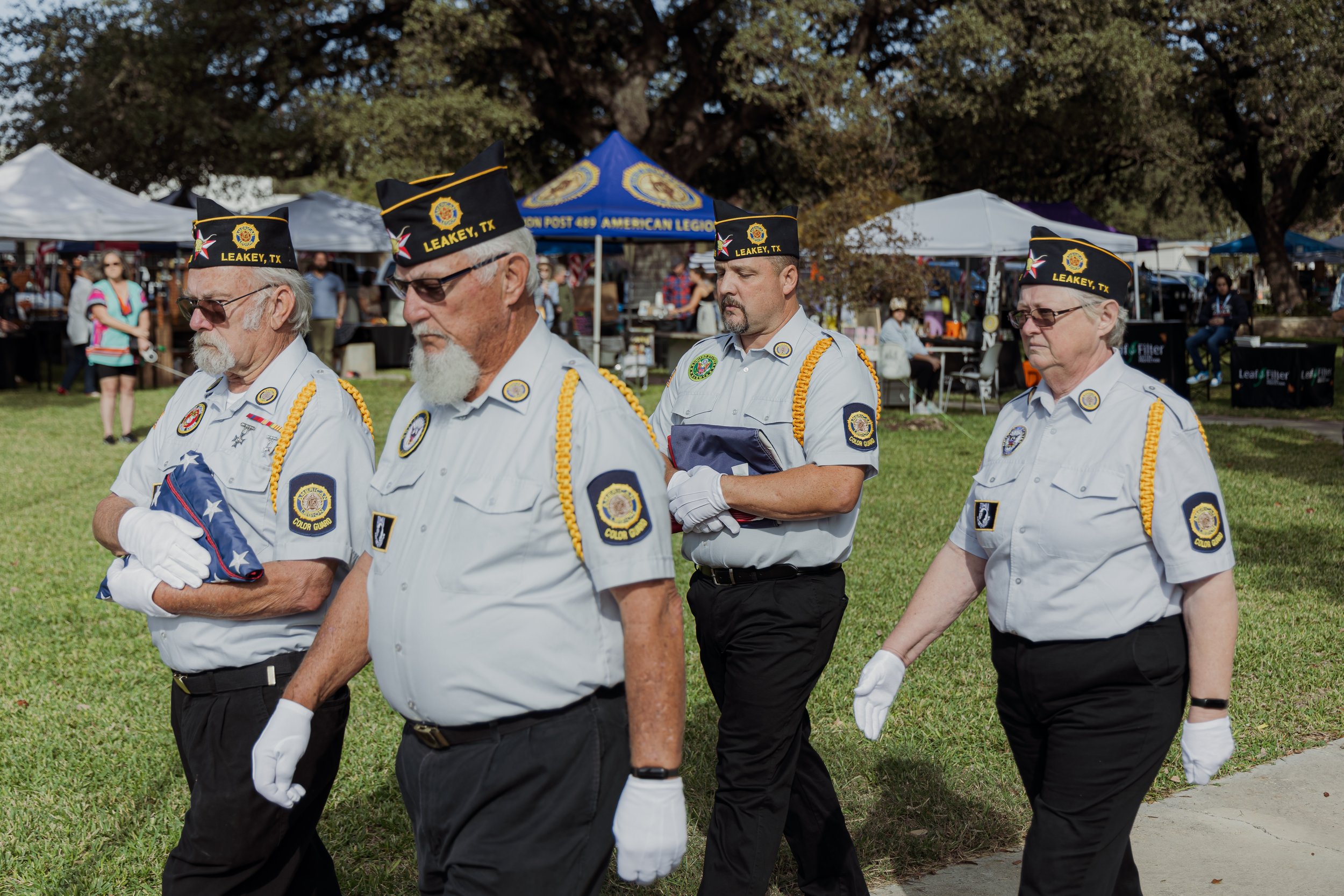 Four veterans wearing uniforms march in a memorial procession, holding a folded American flag. They are outdoors on a grassy area with tents and spectators in the background.