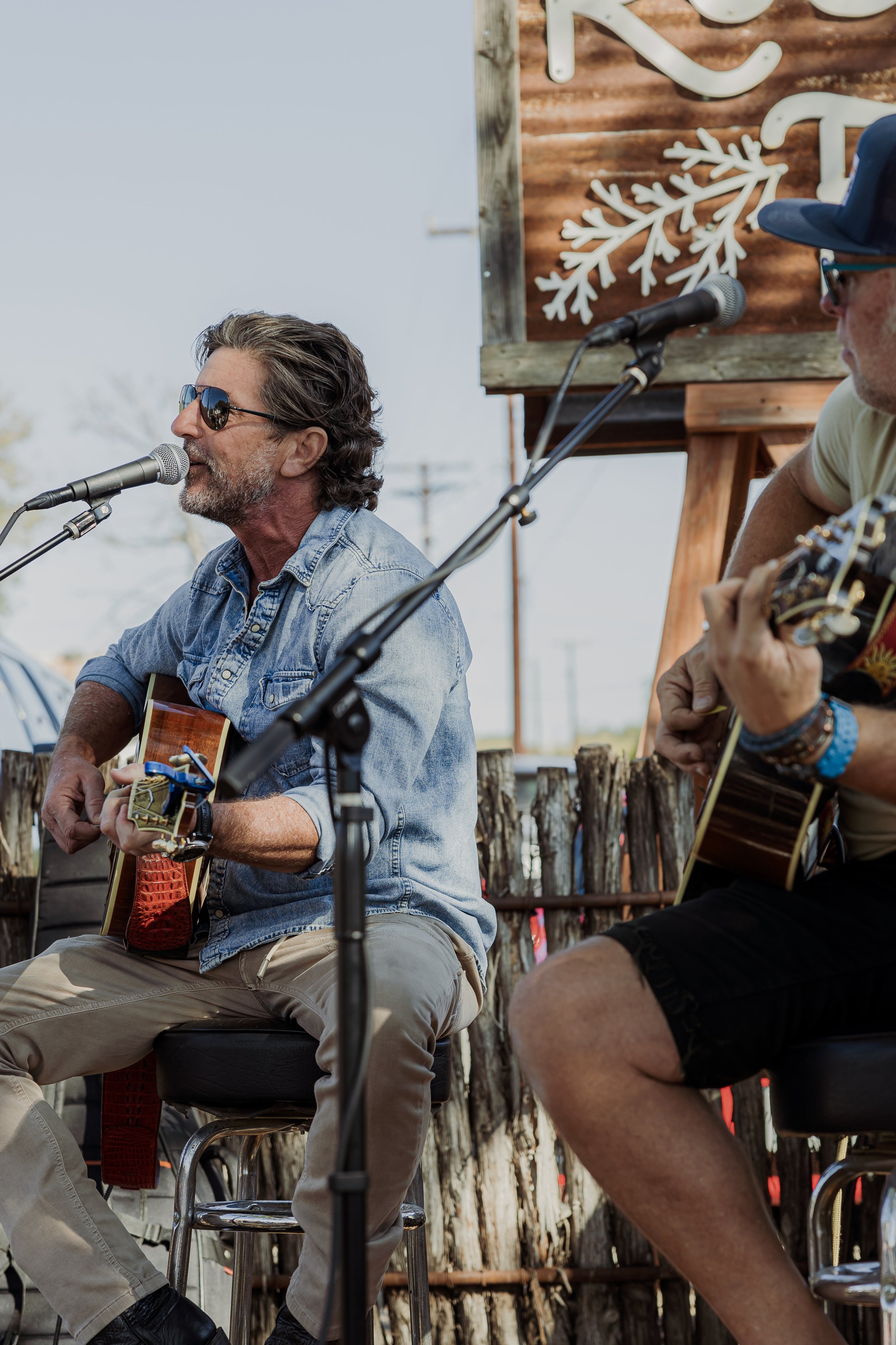 Two men playing guitars and singing into microphones during an outdoor performance, with a wooden sign featuring snowflake and holiday-themed decorations in the background.