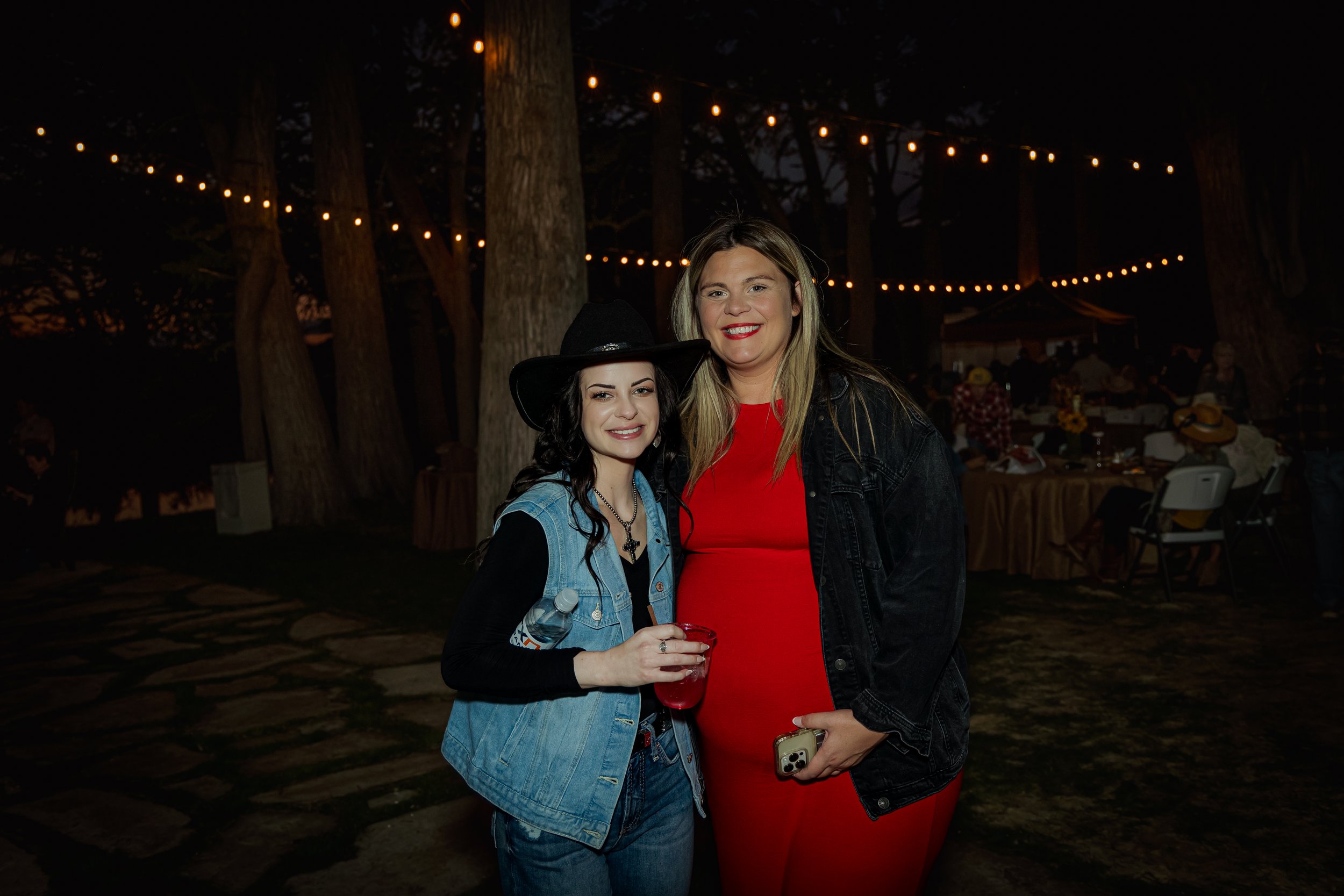Two women standing outdoors at night, smiling, with string lights and trees in the background. One woman is wearing a black hat and denim vest, holding a red cup. The other woman is wearing a red dress and black jacket, holding a phone.