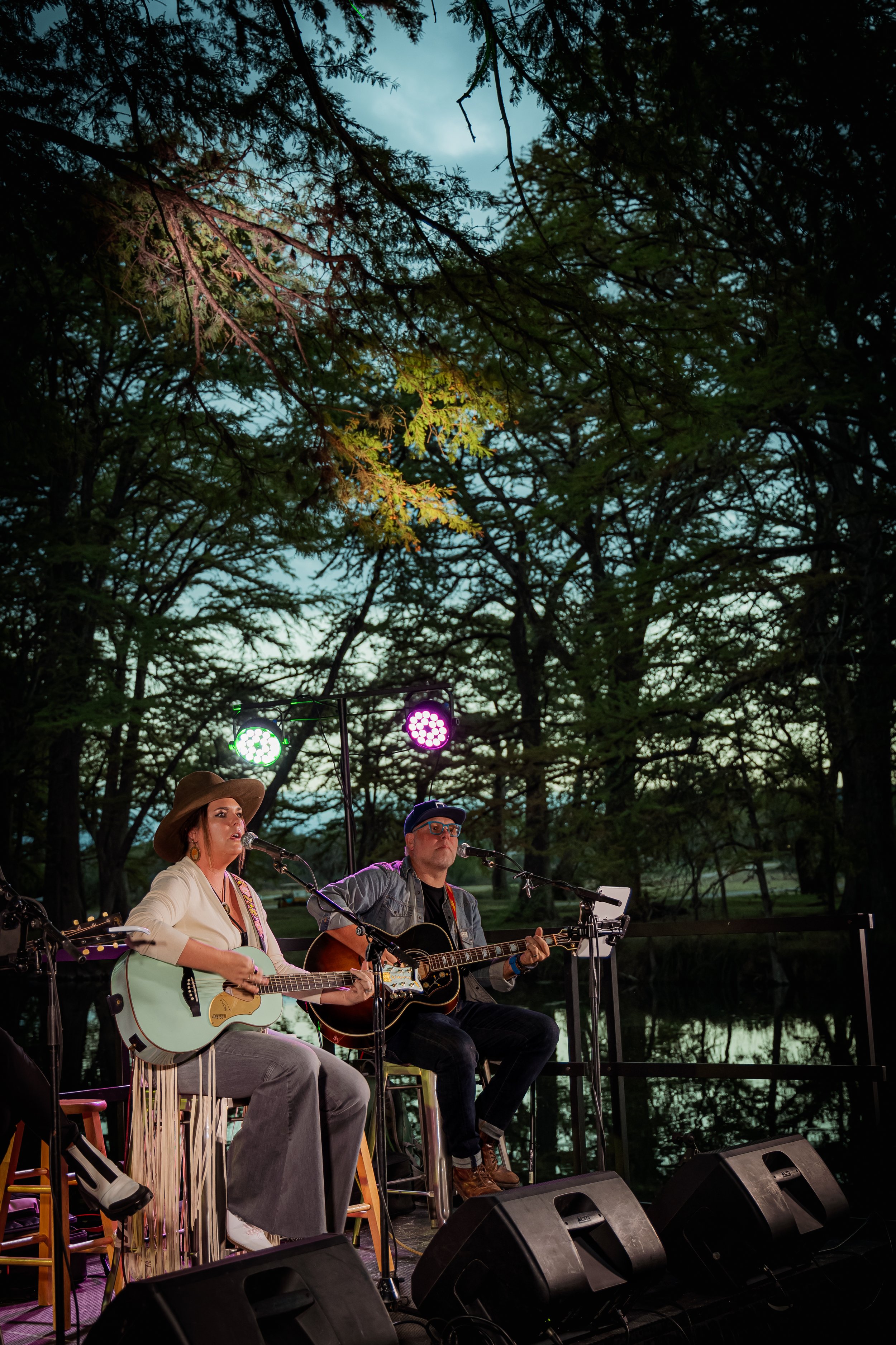 Two musicians performing outdoors at dusk, playing guitars and singing into microphones, surrounded by trees with stage lighting overhead.