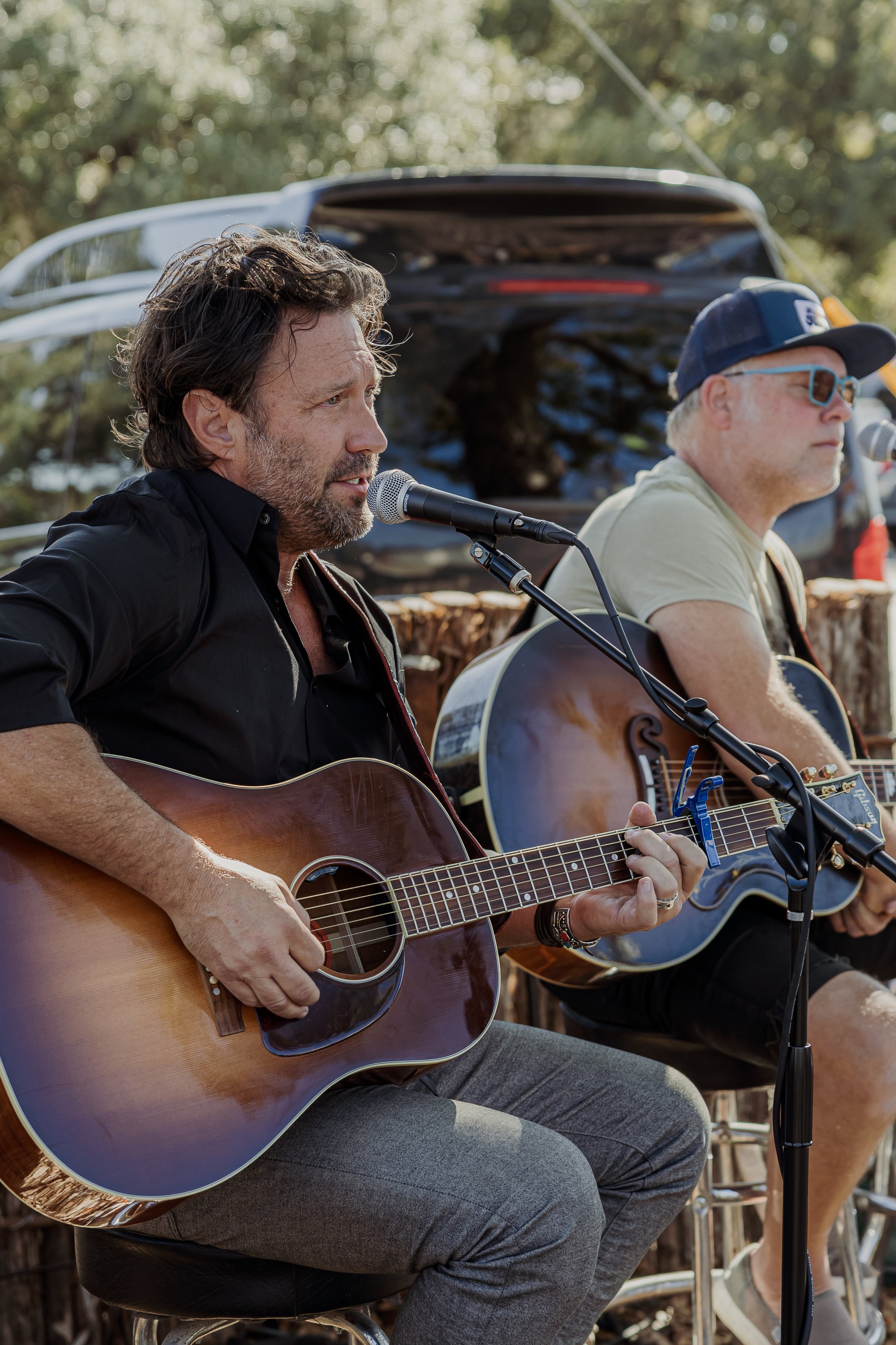 Two men playing acoustic guitars and singing into microphones during outdoor performance