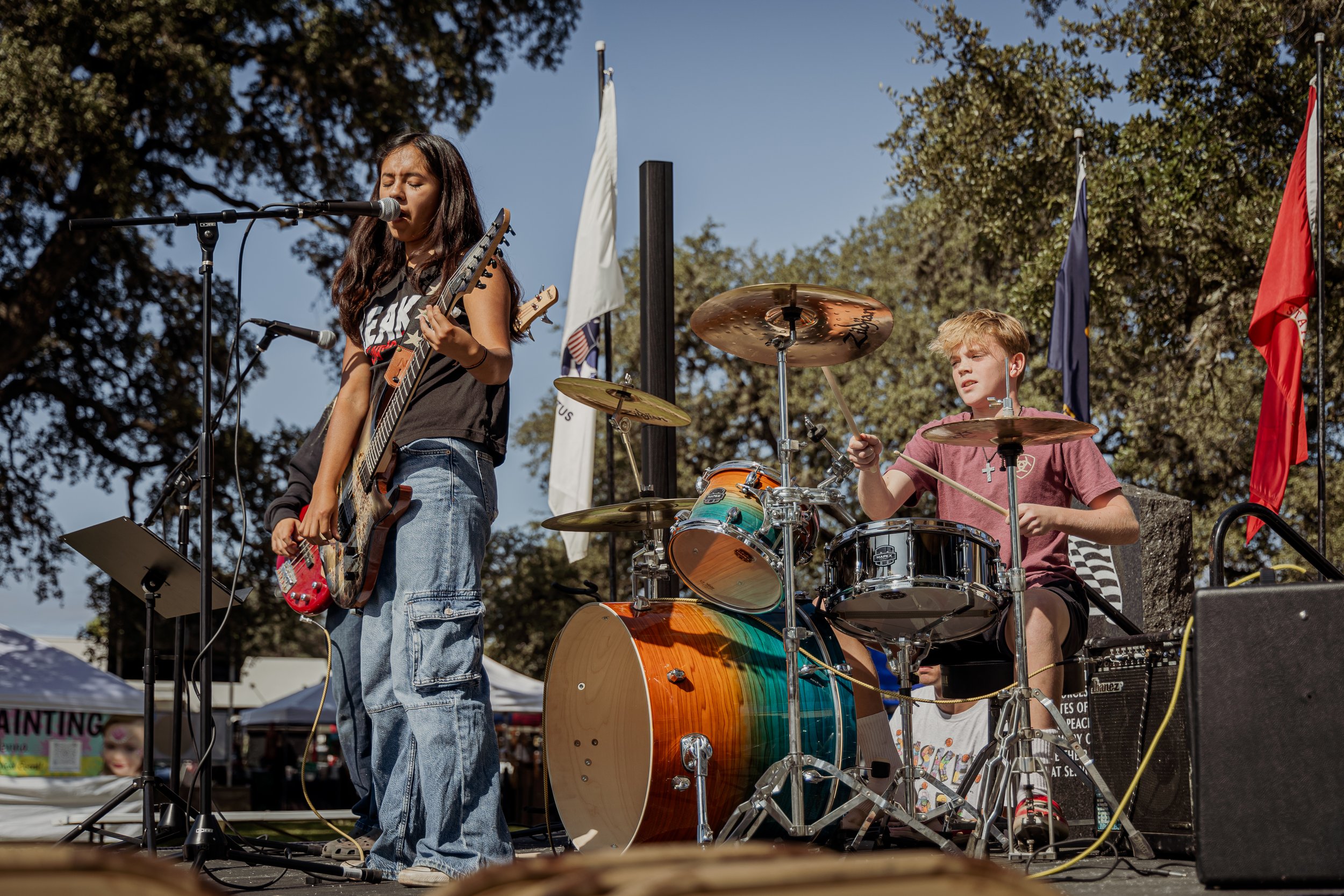 Two young musicians performing outdoors on stage: a girl singing into a microphone while playing an electric guitar, and a boy playing the drums. Background includes trees, flags, and tents.