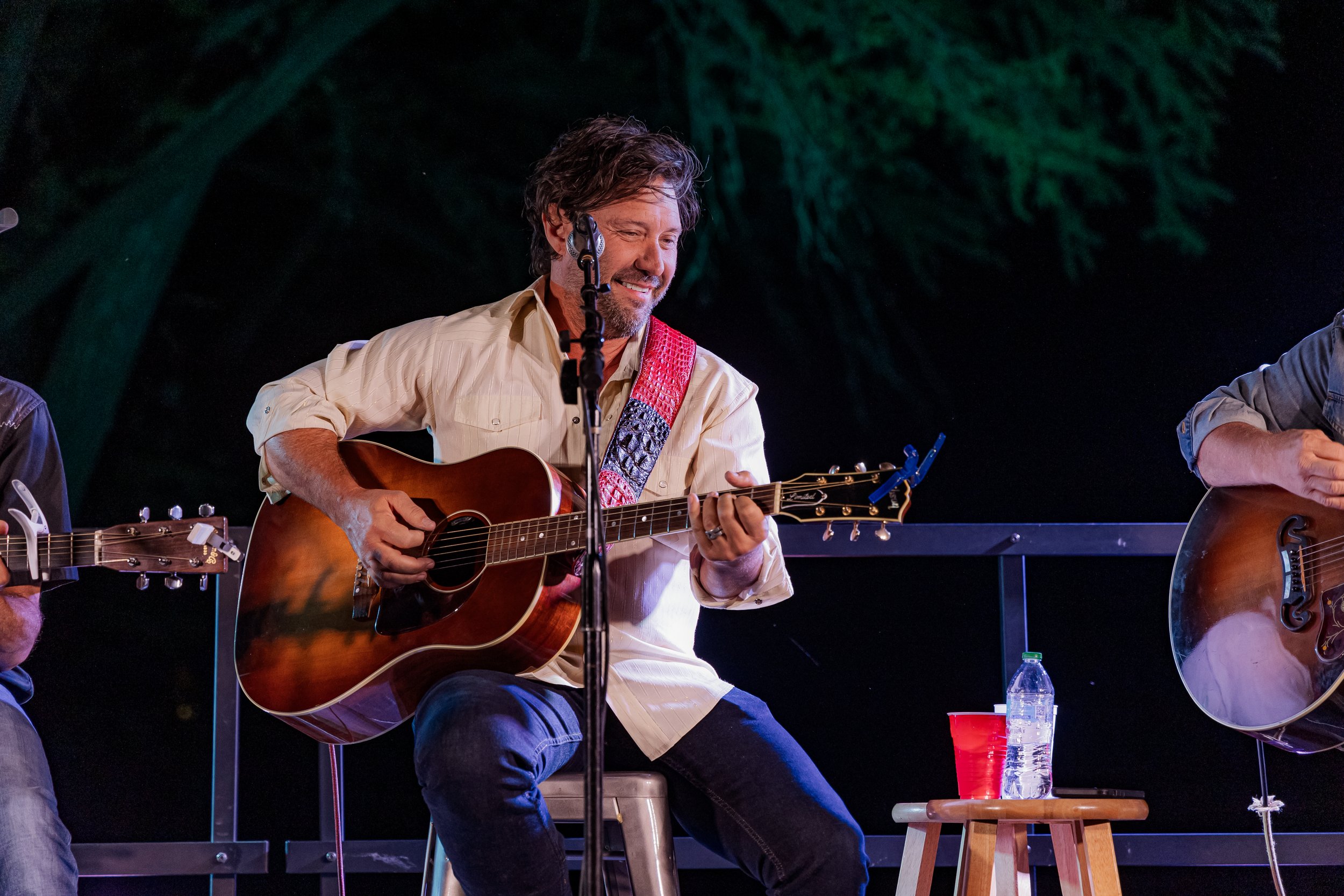 A man with a beard and brown hair playing an acoustic guitar on stage, smiling, with a yellow shirt and a colorful strap. Another musician is partially visible holding a guitar. The background is dark with greenish trees or foliage.