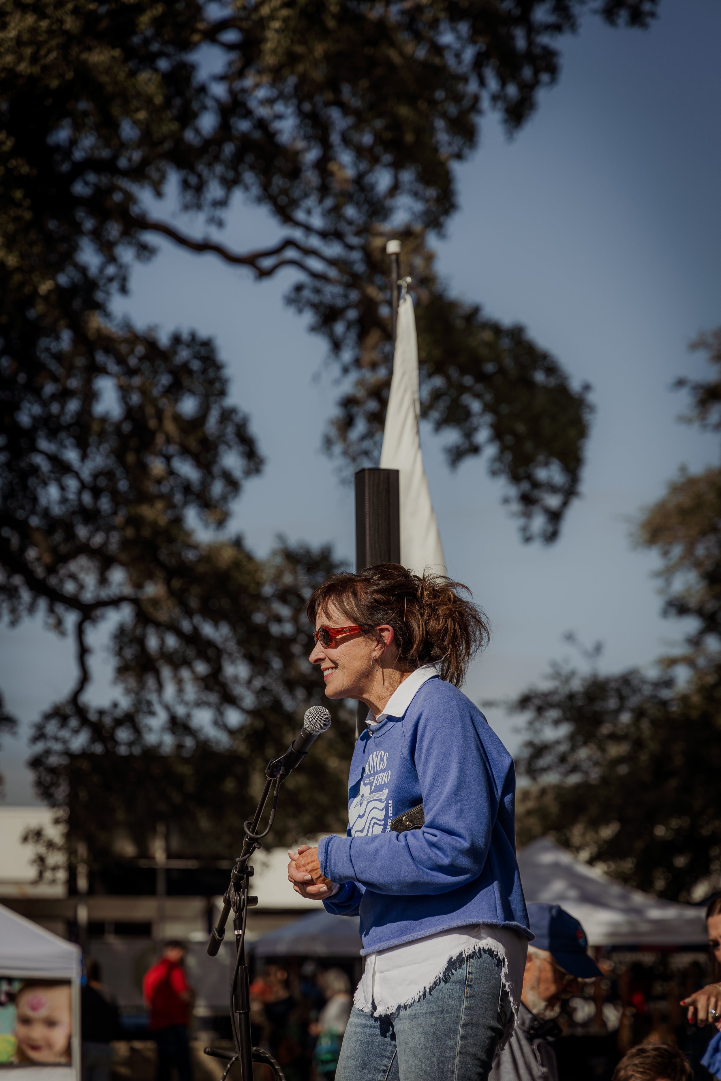 A woman with brown hair and sunglasses speaking into a microphone at an outdoor event, wearing a blue jacket and smiling, with trees and tents in the background.