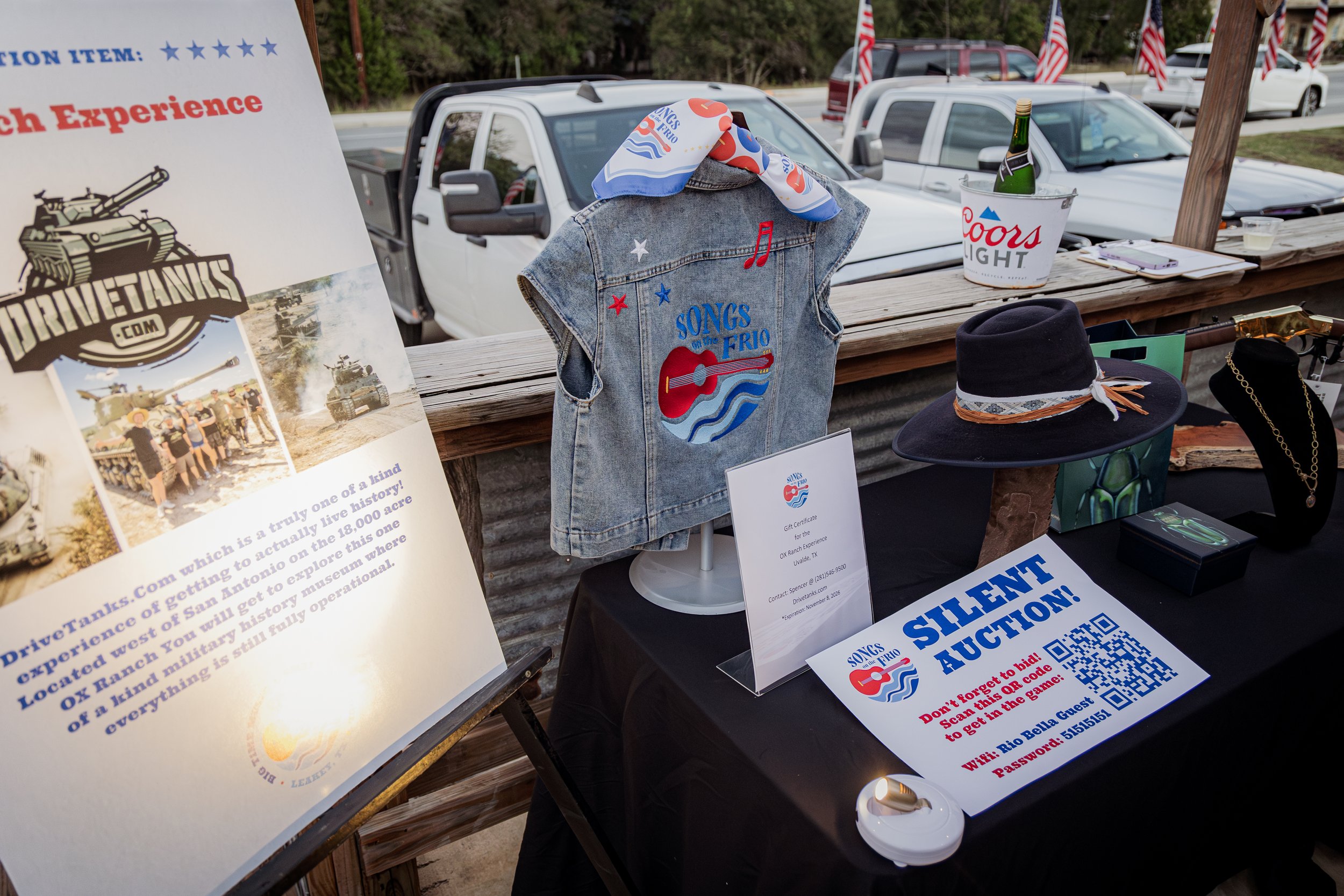 Auction table with a denim vest, hat, jewelry, and a sign for a silent auction. Poster describes a military history museum and the auction includes a QR code with a password.