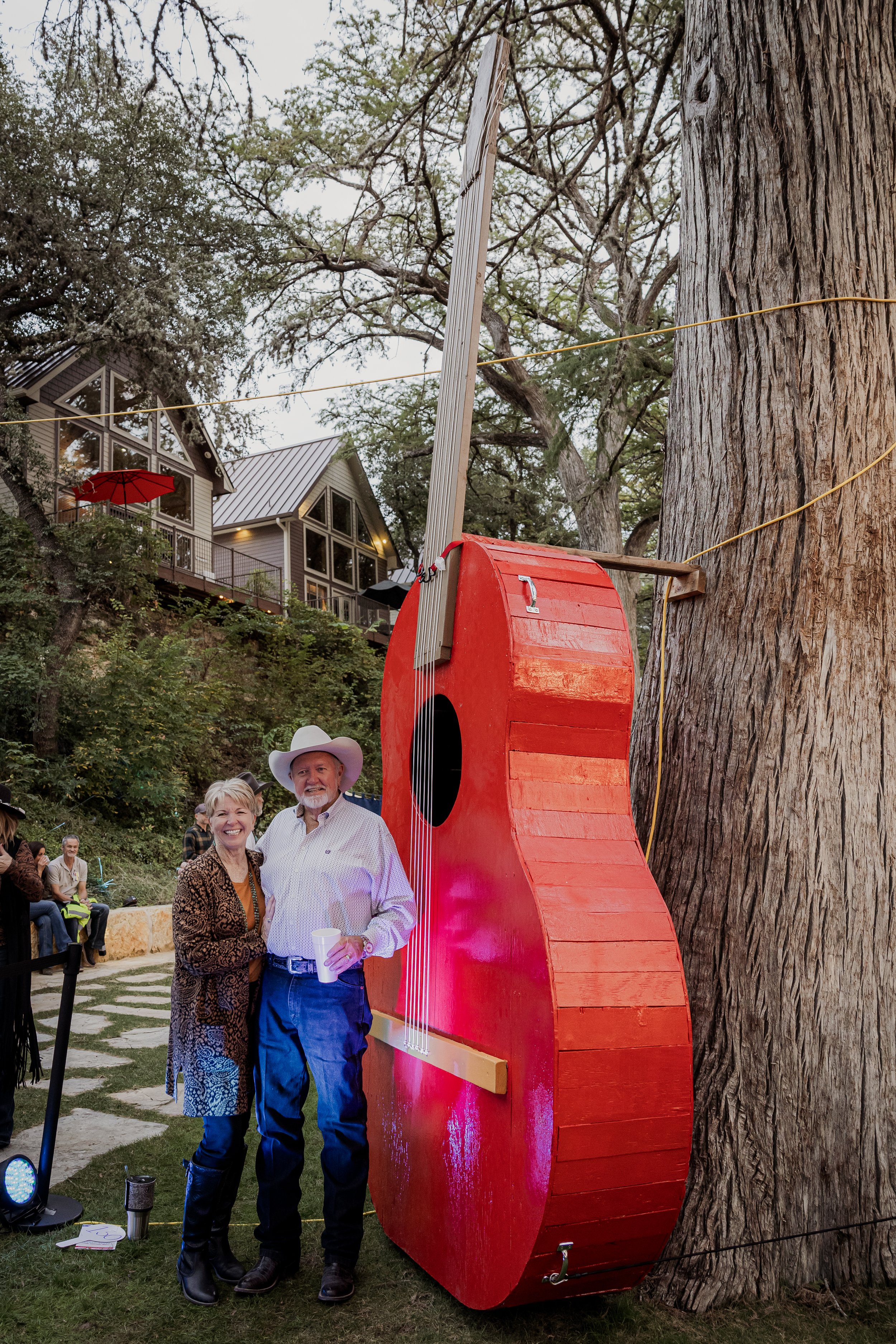 Two people smiling and standing next to a large red guitar-shaped structure attached to a tree during an outdoor event. The man wears a cowboy hat and the woman is dressed in a patterned coat. In the background, there are additional people seated and