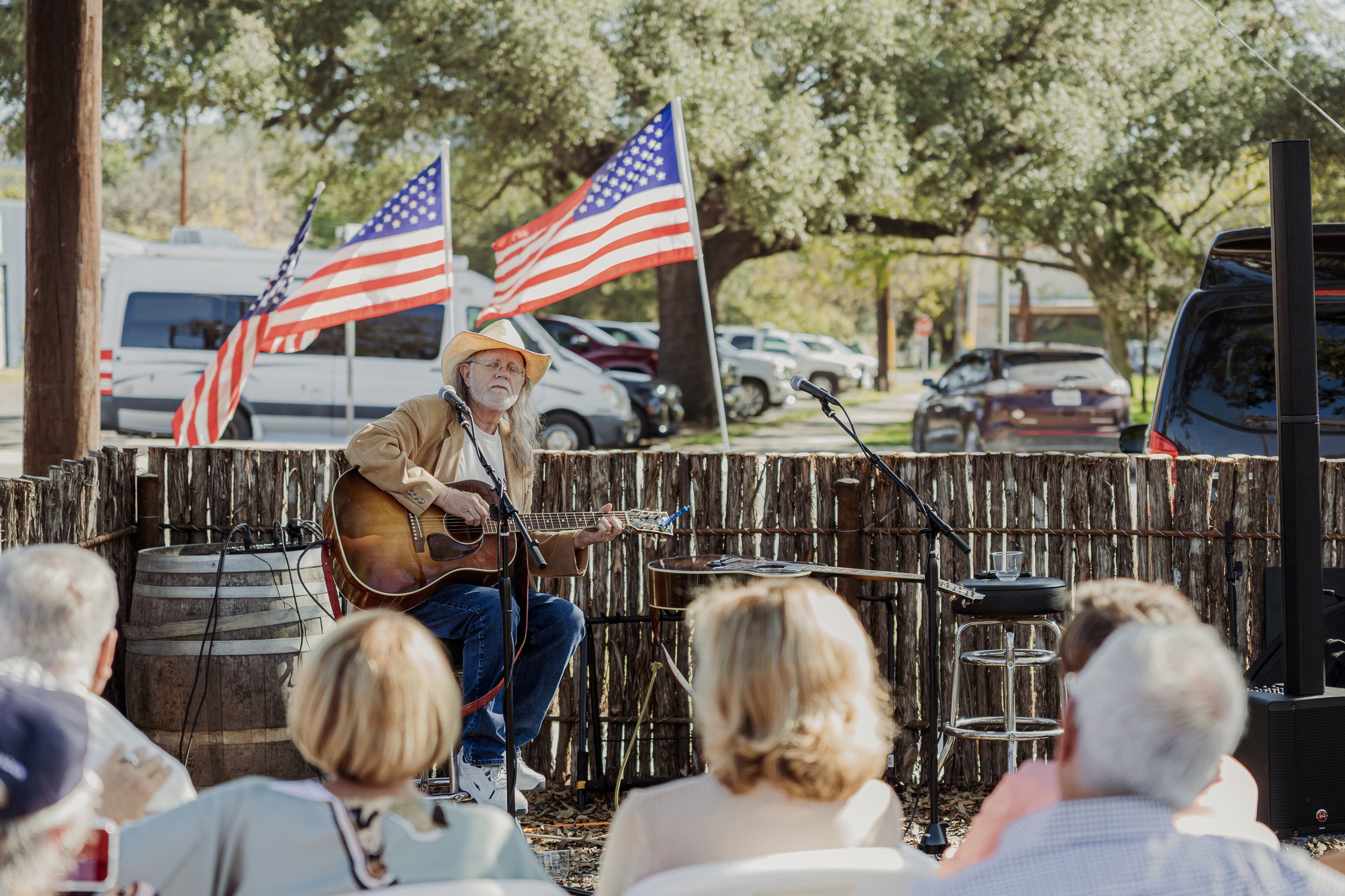A man wearing a cowboy hat plays guitar and sings on a small outdoor stage, with American flags behind him and audience members seated in front, under tree shade.