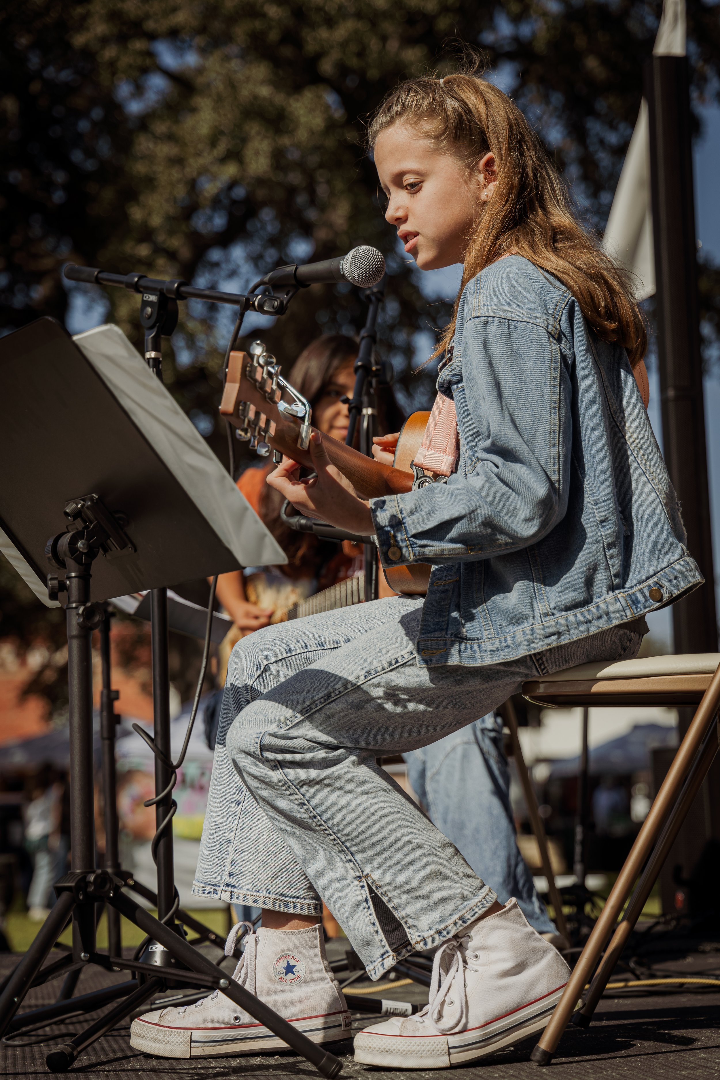 A young girl playing acoustic guitar and singing into a microphone during an outdoor performance, with another girl visible in the background.