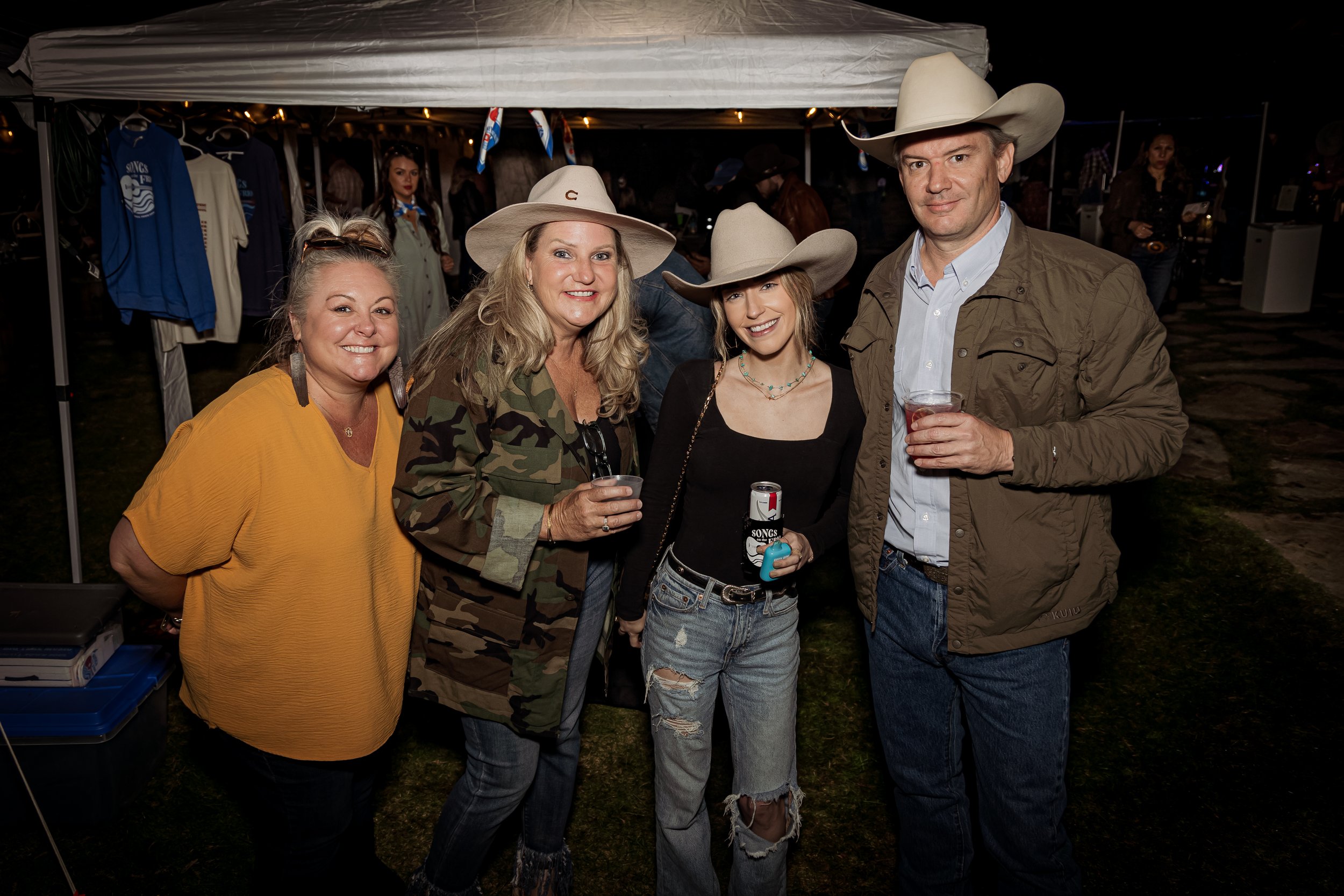 Four people standing together at an outdoor event at night, wearing cowboy hats, with a tent and vendor booths visible in the background, holding drinks and smiling at the camera.