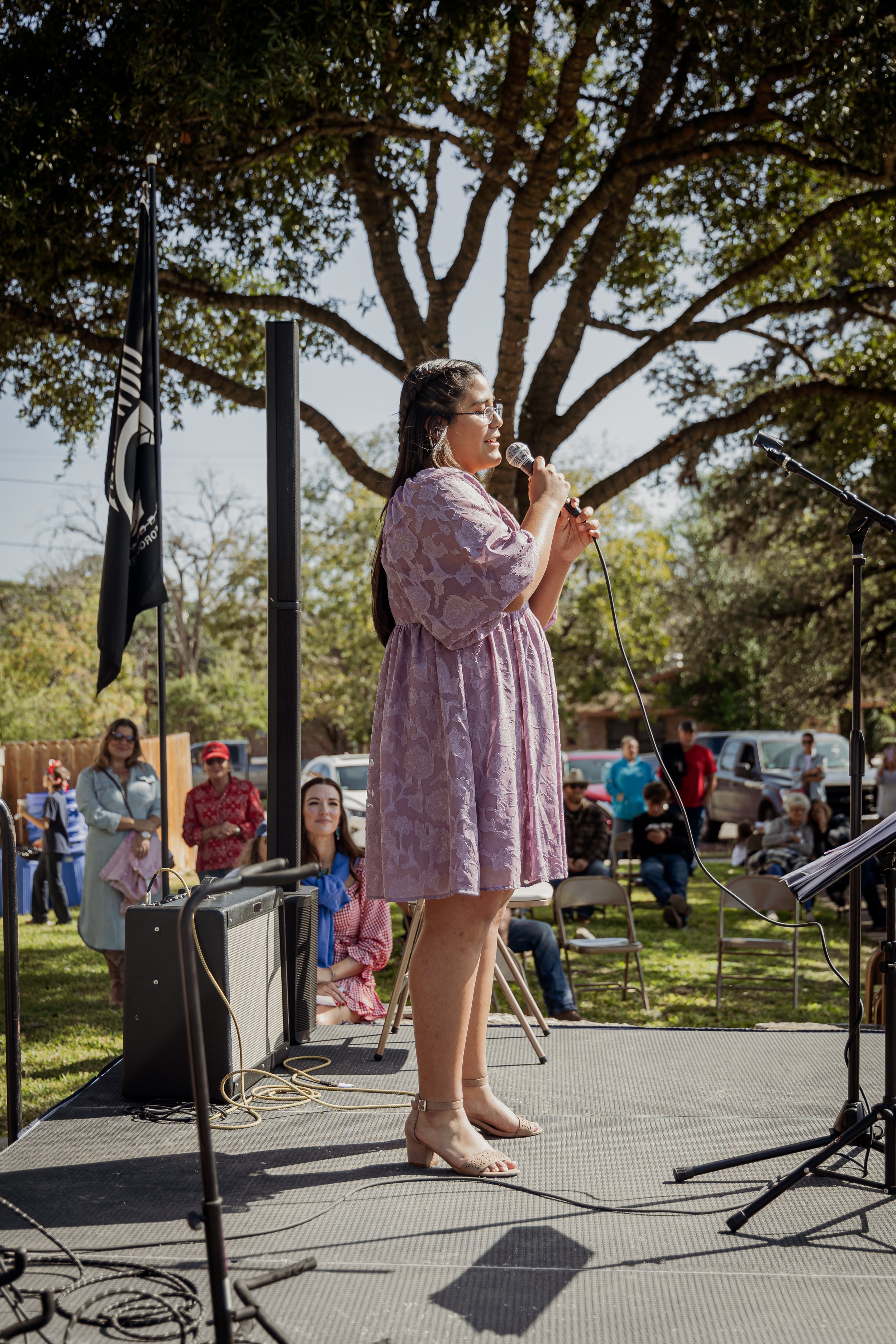 Woman in pink dress with glasses speaking into microphone on outdoor stage, audience seated and watching, large tree in background with sunny sky.