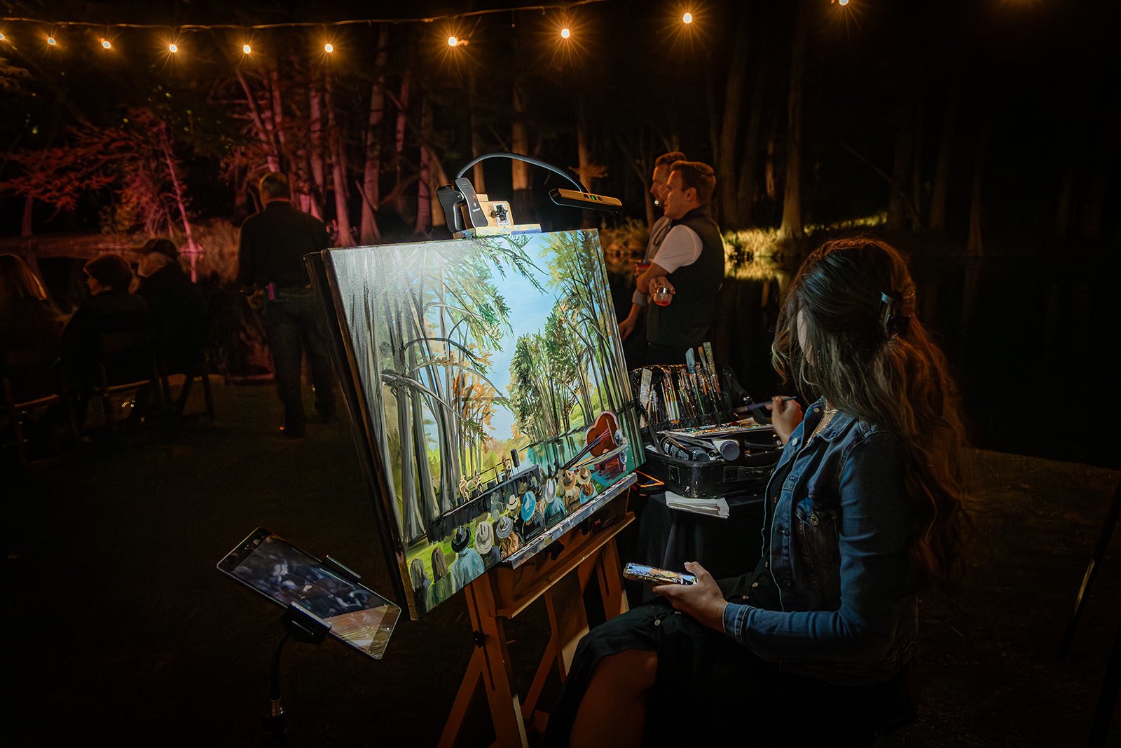 An outdoor nighttime scene of a woman sitting at an easel painting a landscape of a forest and a lake. There is an artist's case and painting supplies on a table beside her, with a smartphone mounted on a small stand nearby. In the background, severa