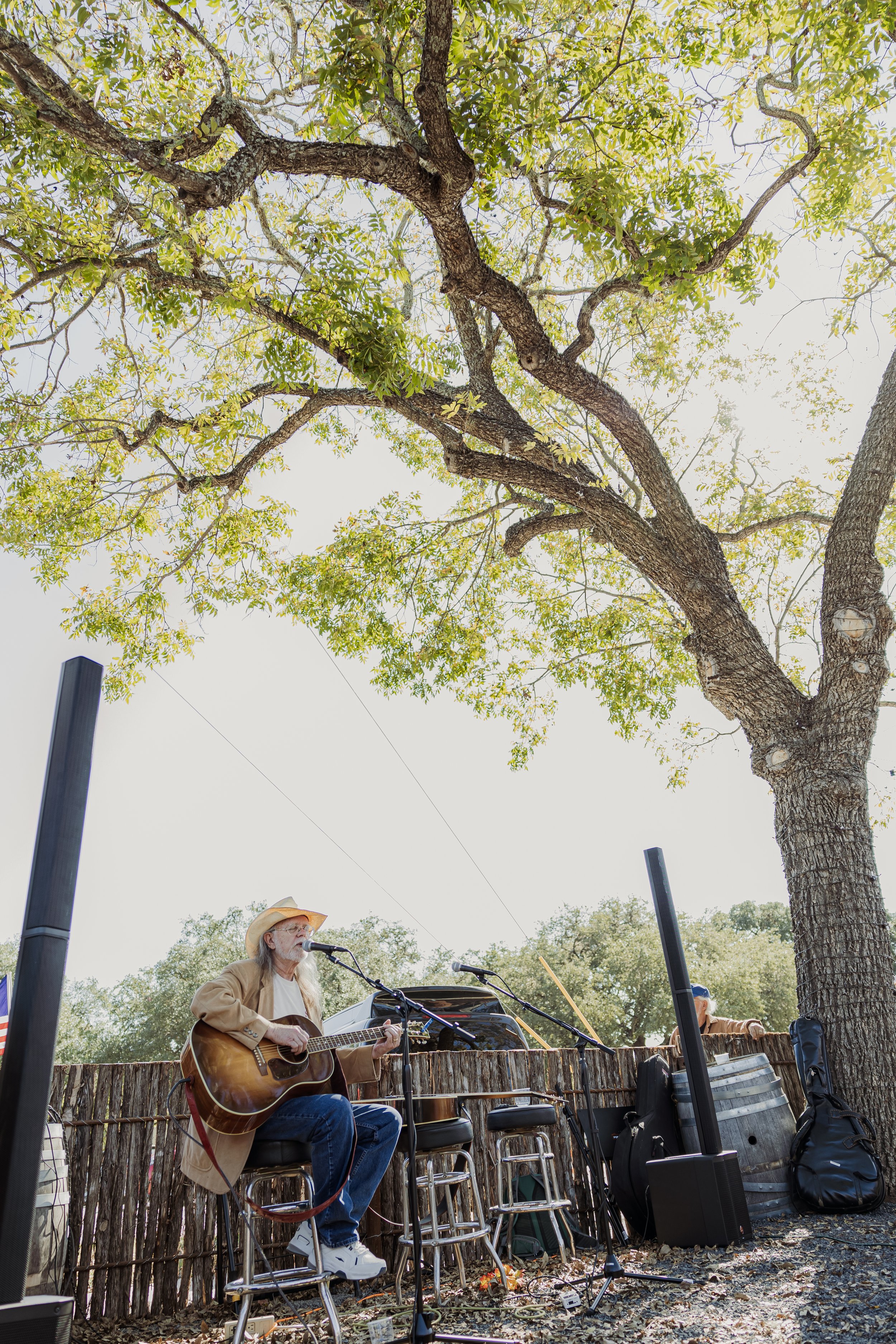 A man with long hair and a beard, wearing a cowboy hat and brown jacket, playing an acoustic guitar and singing into a microphone outdoors. He is sitting on a stool in front of a rustic wooden fence beneath a large tree with green leaves. There are m