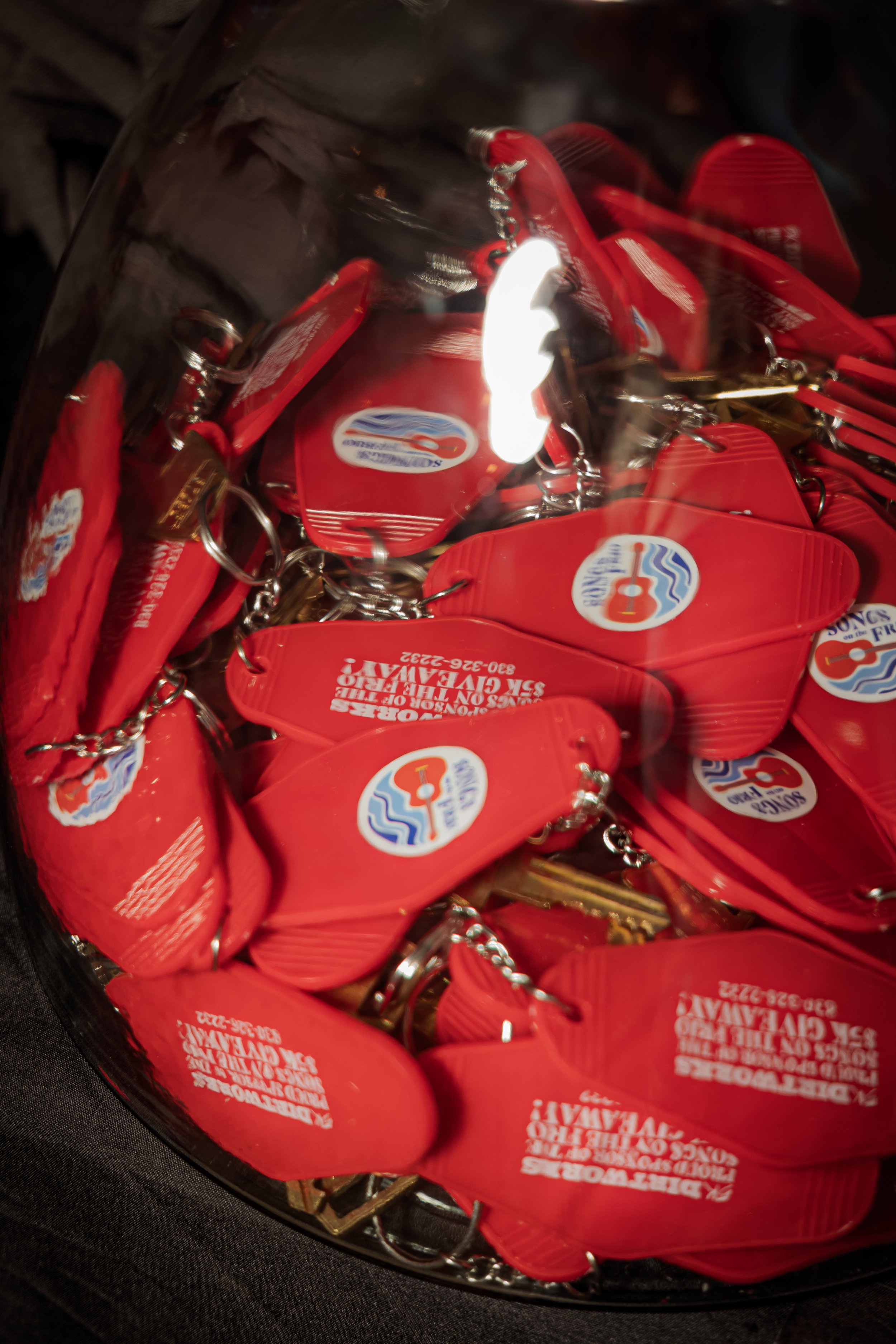 Close-up of red keychains with a logo of a guitar and water, and some printed text, inside a clear container.