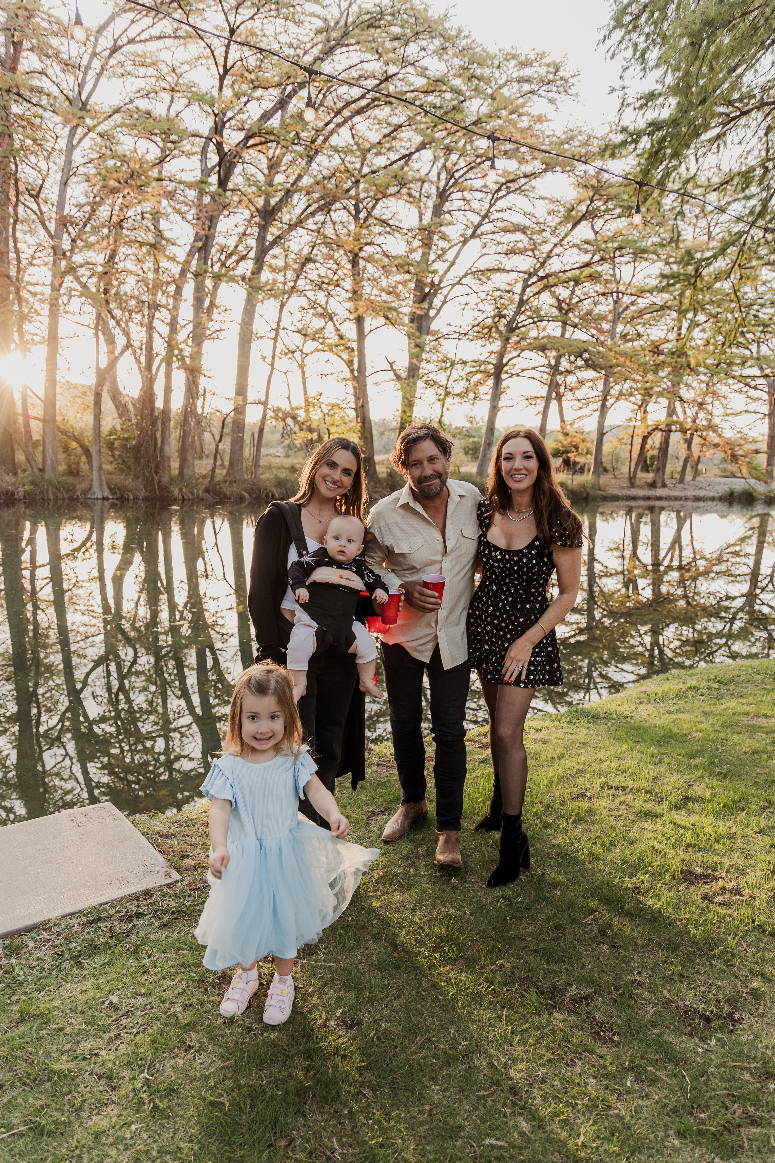 A group of five people, including two women, one man, and two young children, standing outdoors near a body of water with trees in the background during the sunset, smiling at the camera.