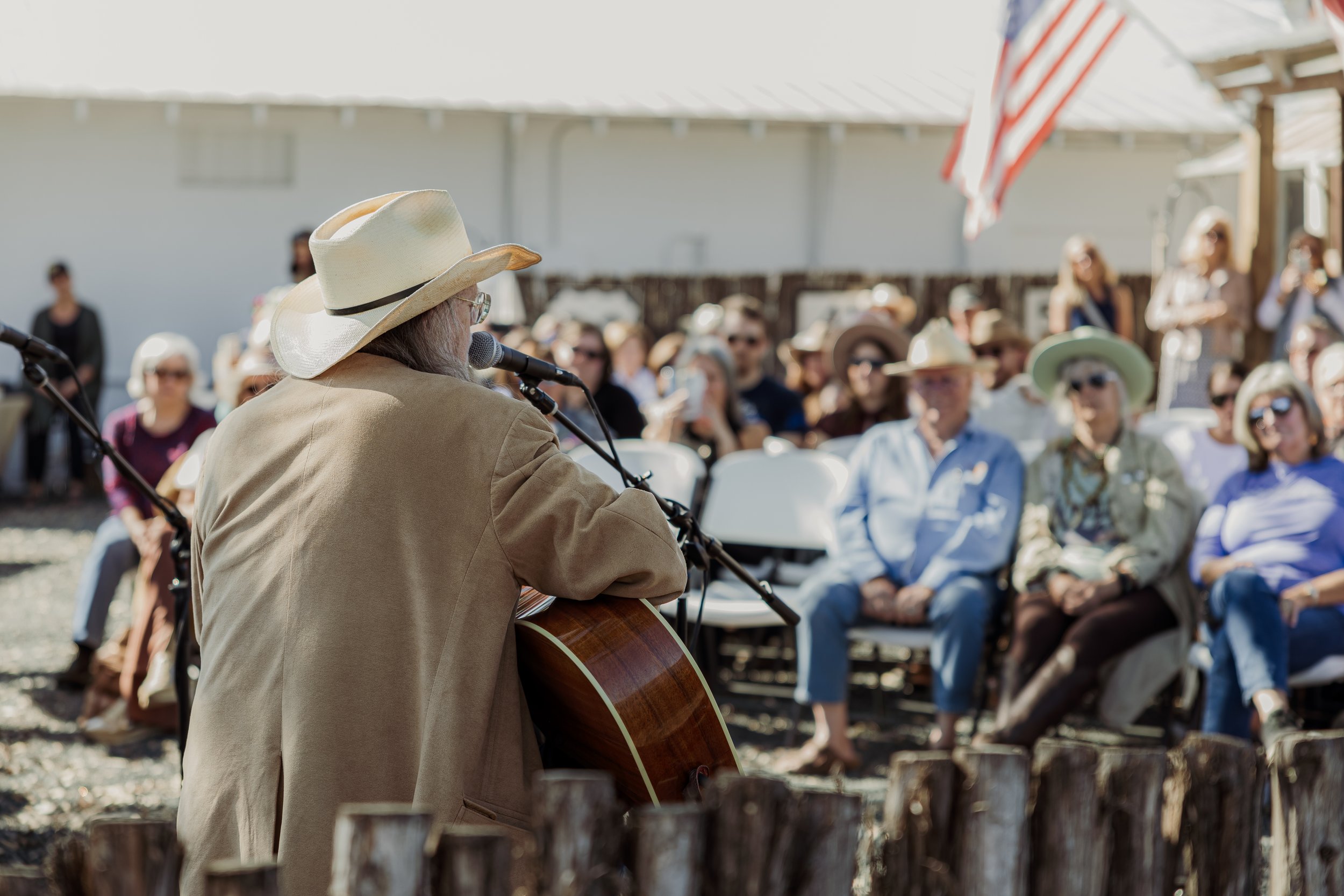 A man wearing a cowboy hat and beige jacket playing guitar and singing into a microphone at an outdoor event with an audience seated in front of him.