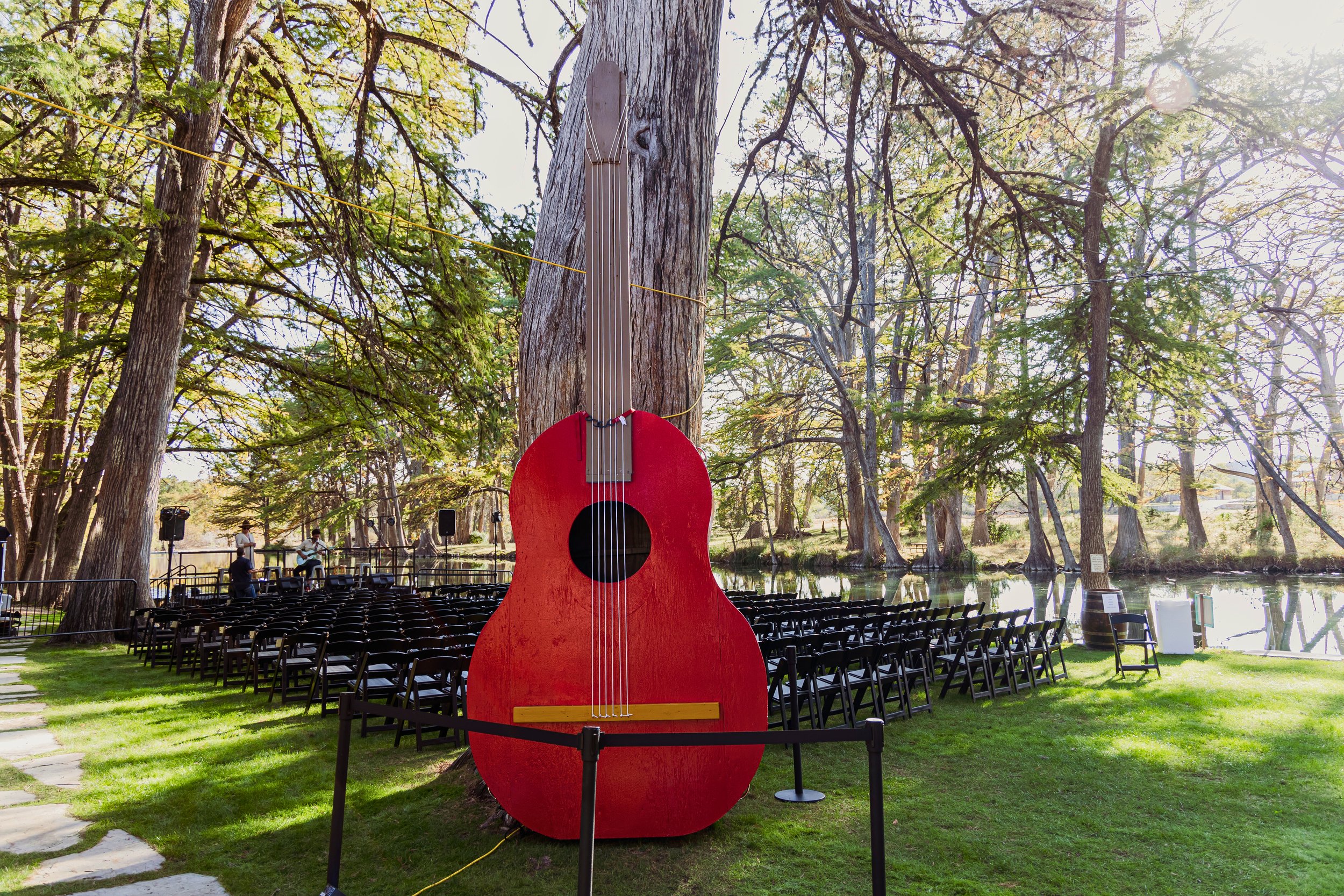 A giant decorative red guitar standing in an outdoor park by a pond, with chairs arranged for an event and musicians setting up in the background, surrounded by tall trees.