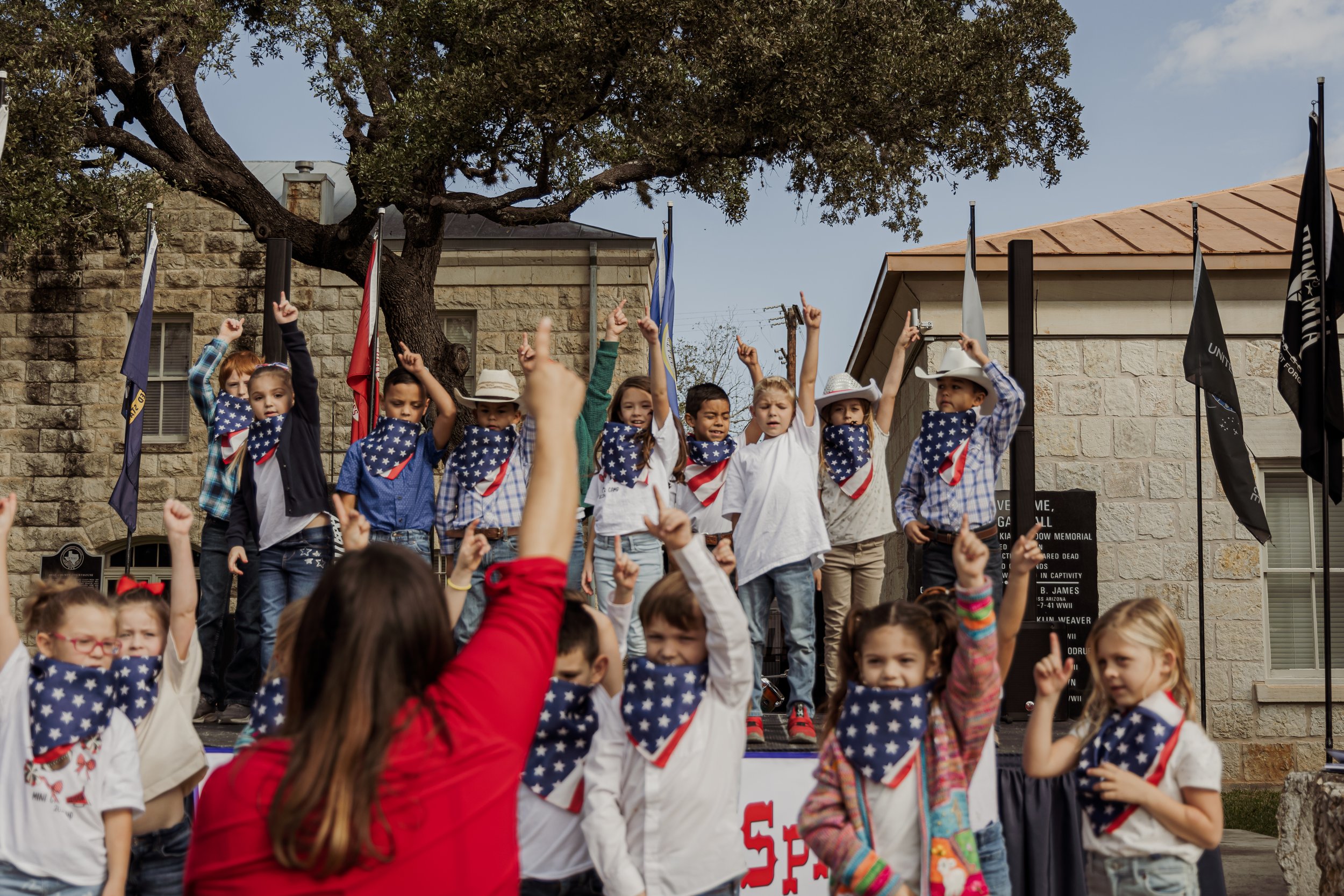 Children participating in a patriotic event, wearing bandanas with American flag patterns, standing on a stage and raising their hands, with adults and flags in the background.