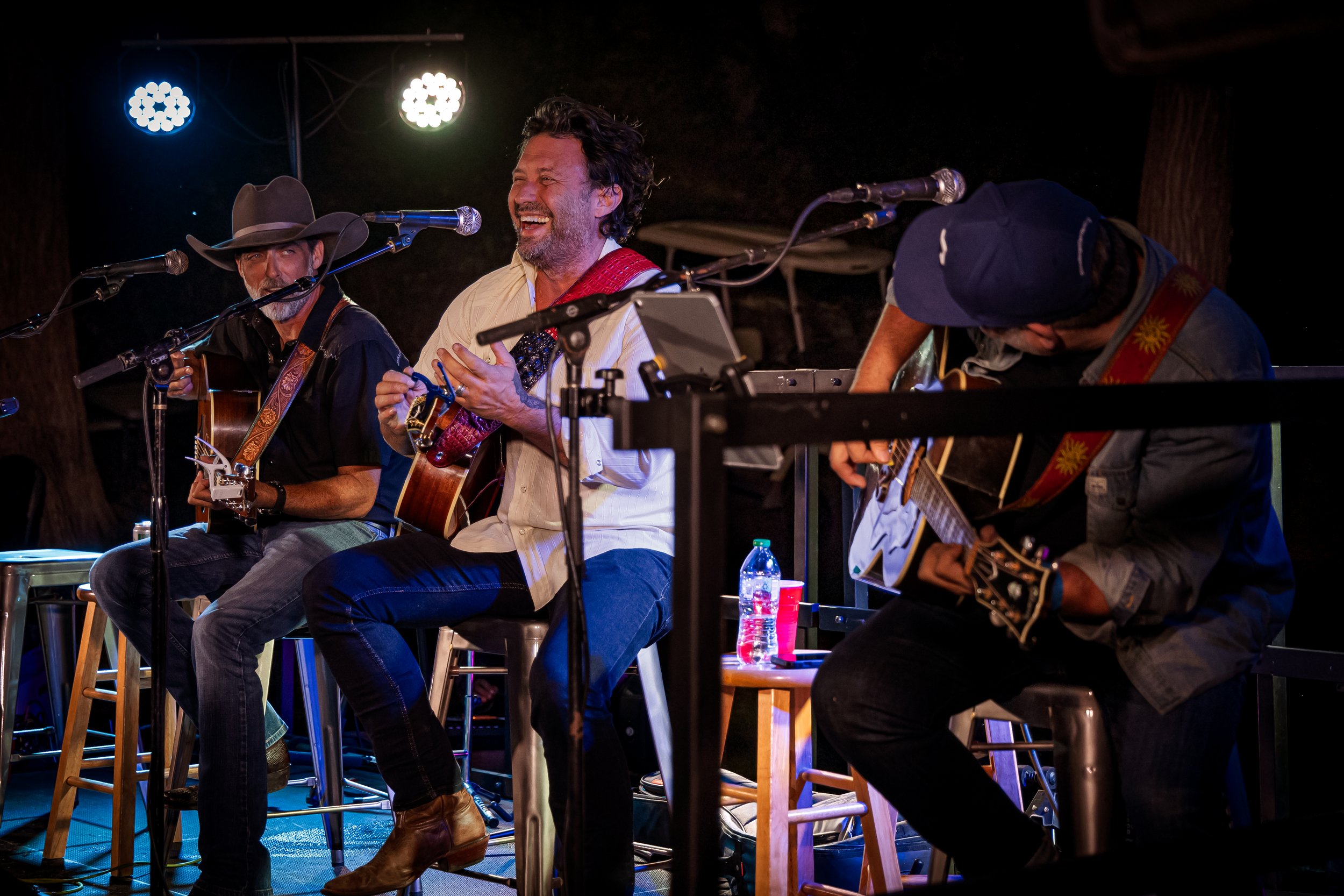 Three male musicians performing with guitars on a stage, one singing, with stage lights overhead.