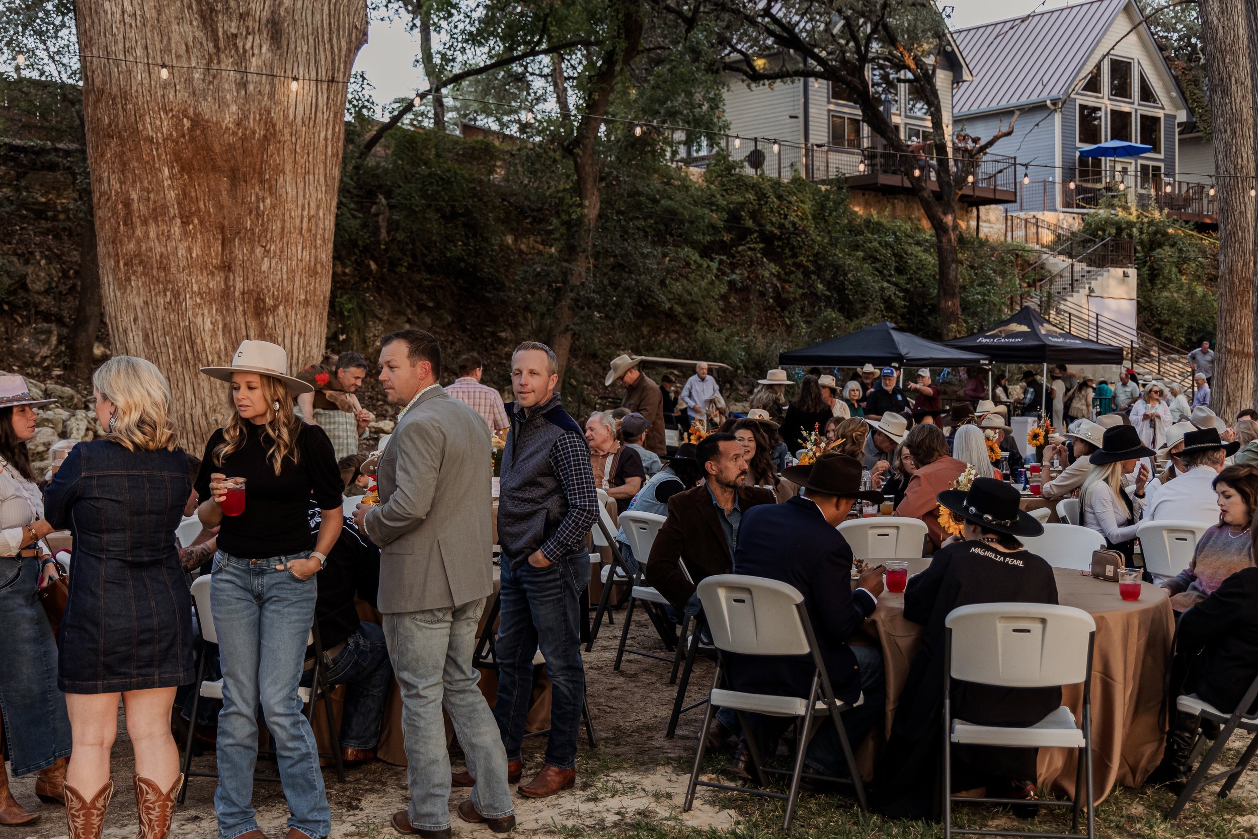 Outdoor social gathering with people seated at round tables and standing, some wearing hats, in a backyard with large trees, string lights, and a house with a deck in the background.