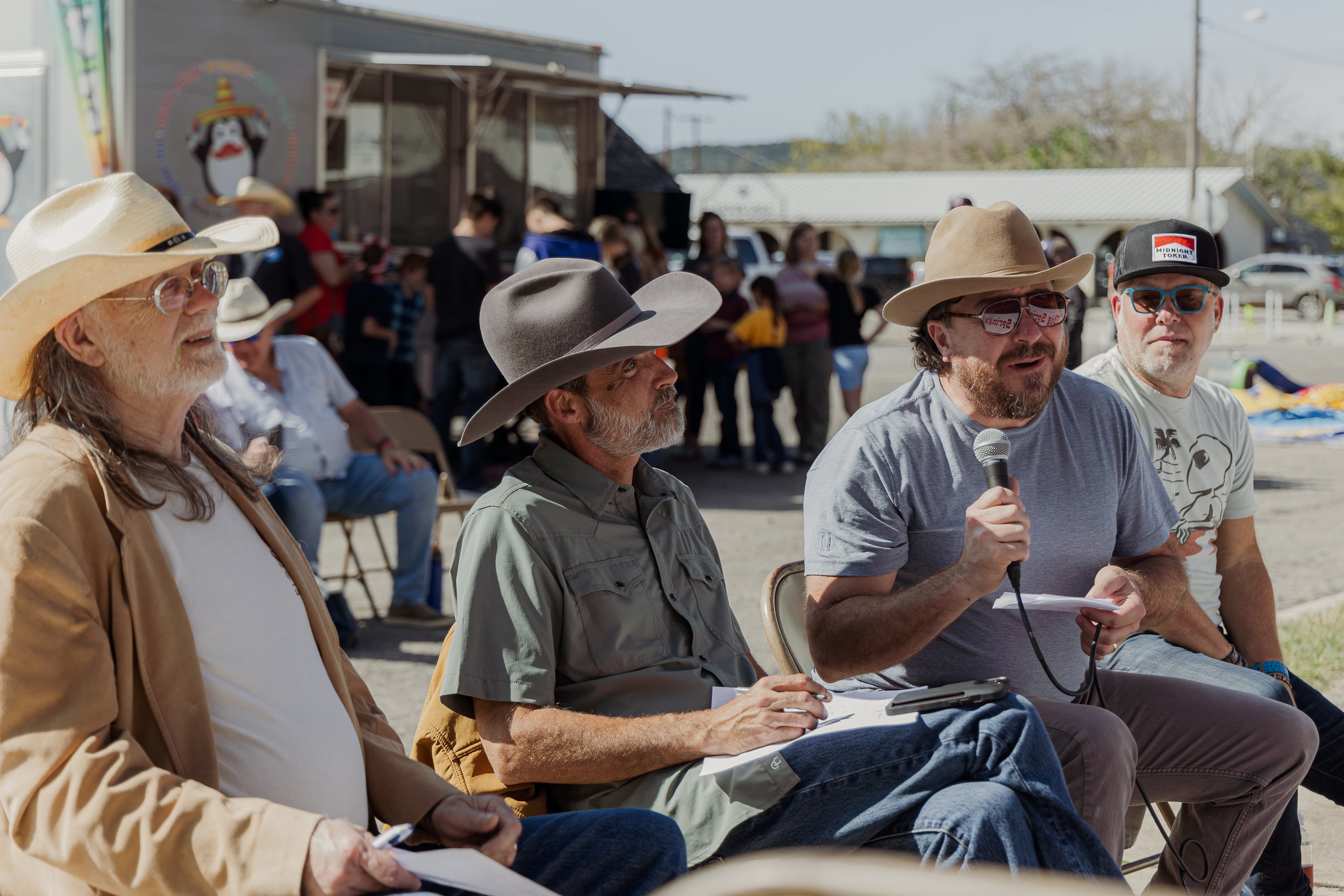 Four men sitting outdoors at a panel, with one speaking into a microphone. All wear hats and sunglasses, and there is a crowd in the background.